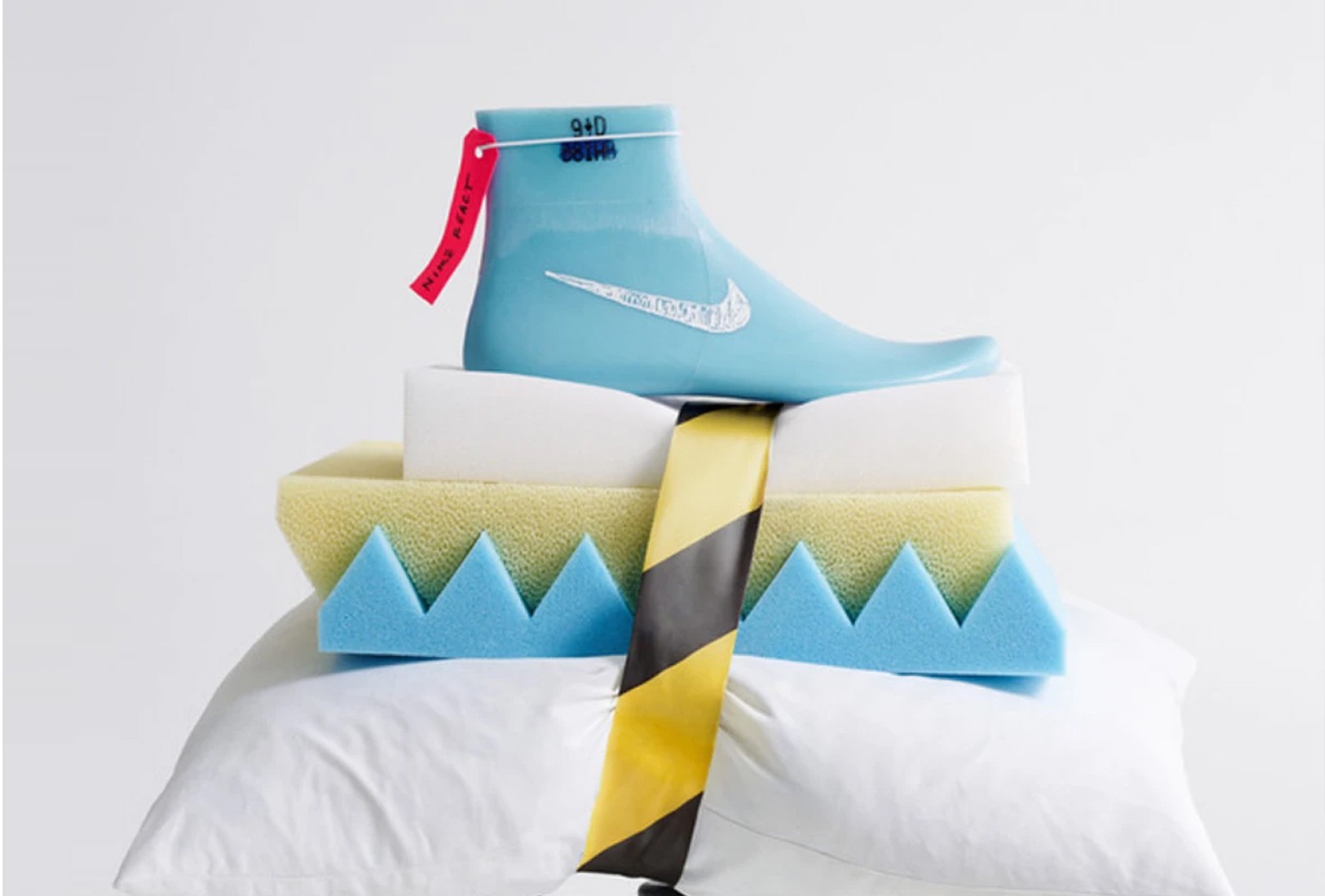 A blue shoe model with a Nike logo and red tag rests on stacked foam pieces and a white pillow, with a yellow and black striped ribbon draped across.
