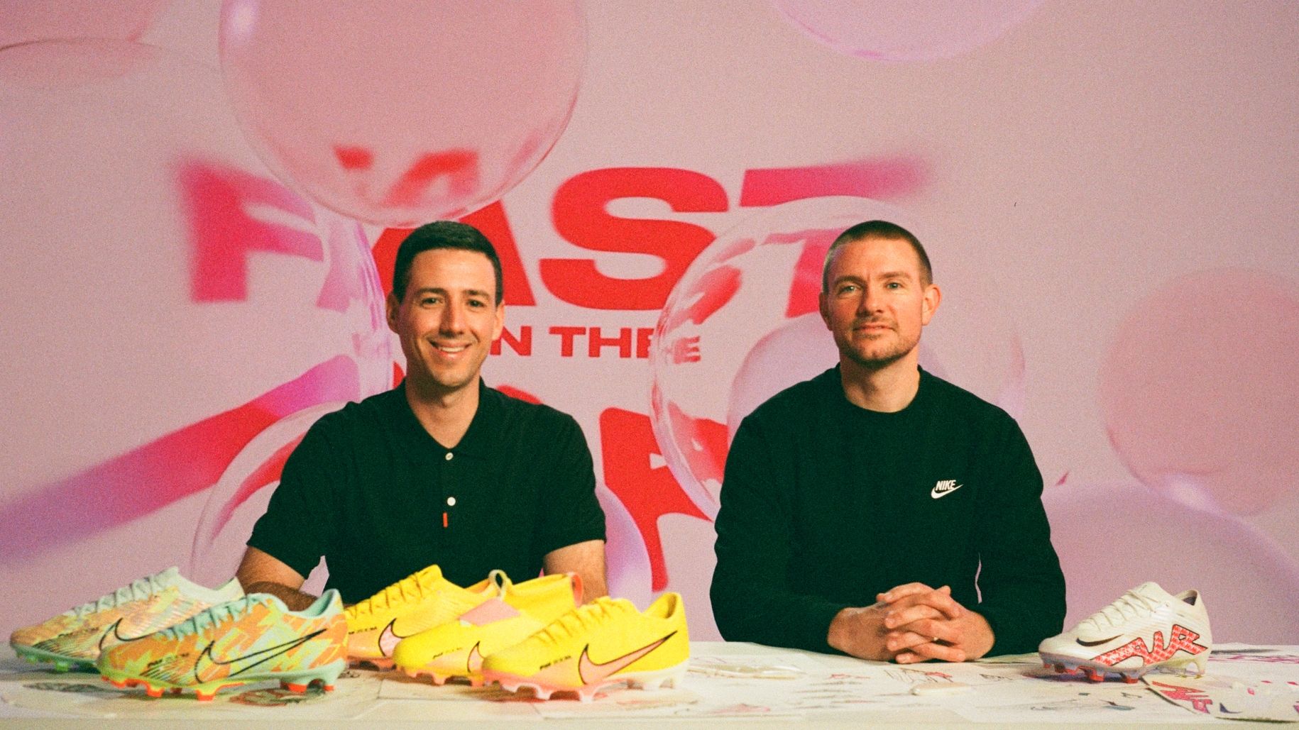 Two event hosts sitting at desk with Nike Mercurial boots in all colorways on desk. Fast is in the air with Bubbles is the background