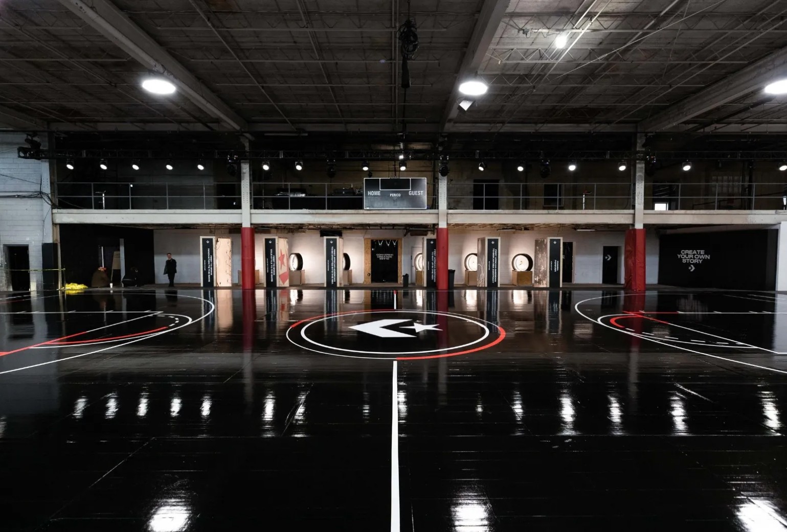 A glossy indoor basketball court with a black floor, white and red lines, and a Converse logo at center, inside an industrial-style building.