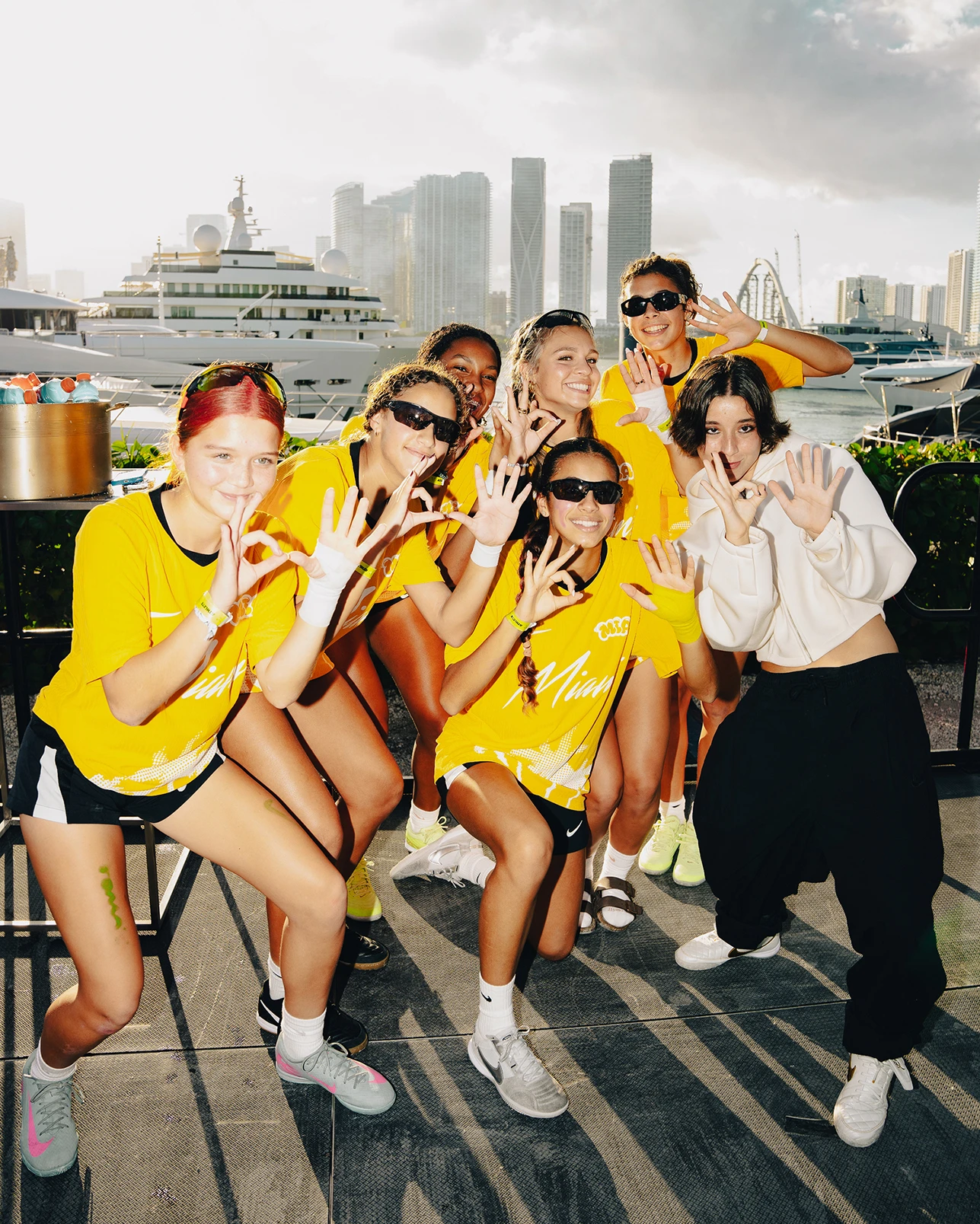 A group of six people in yellow shirts and one person in white pose together outdoors near yachts and skyscrapers, some making hand signs and smiling at the camera.