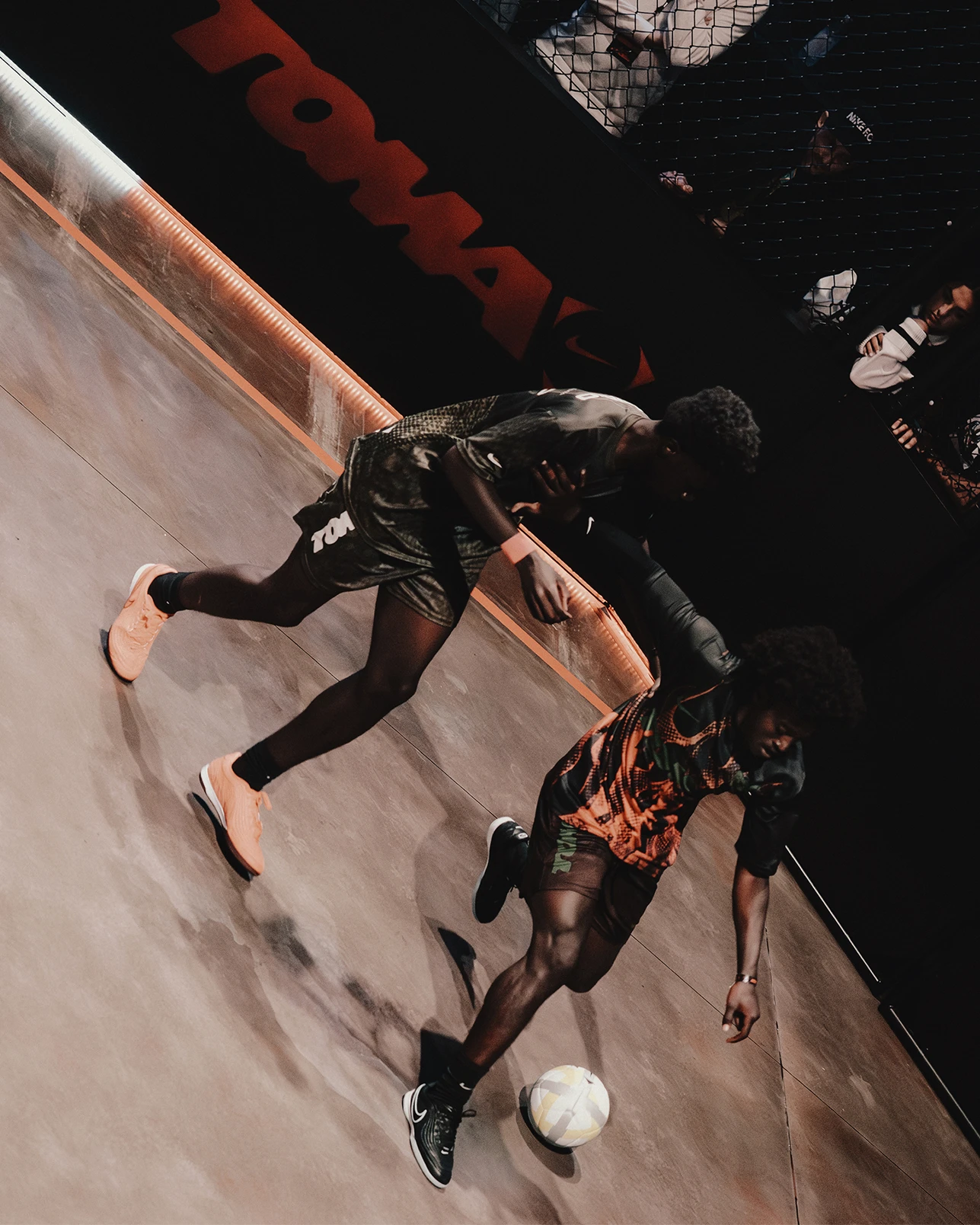 Two people play indoor soccer on a court, competing for the ball. A black net separates the players from spectators in the background.