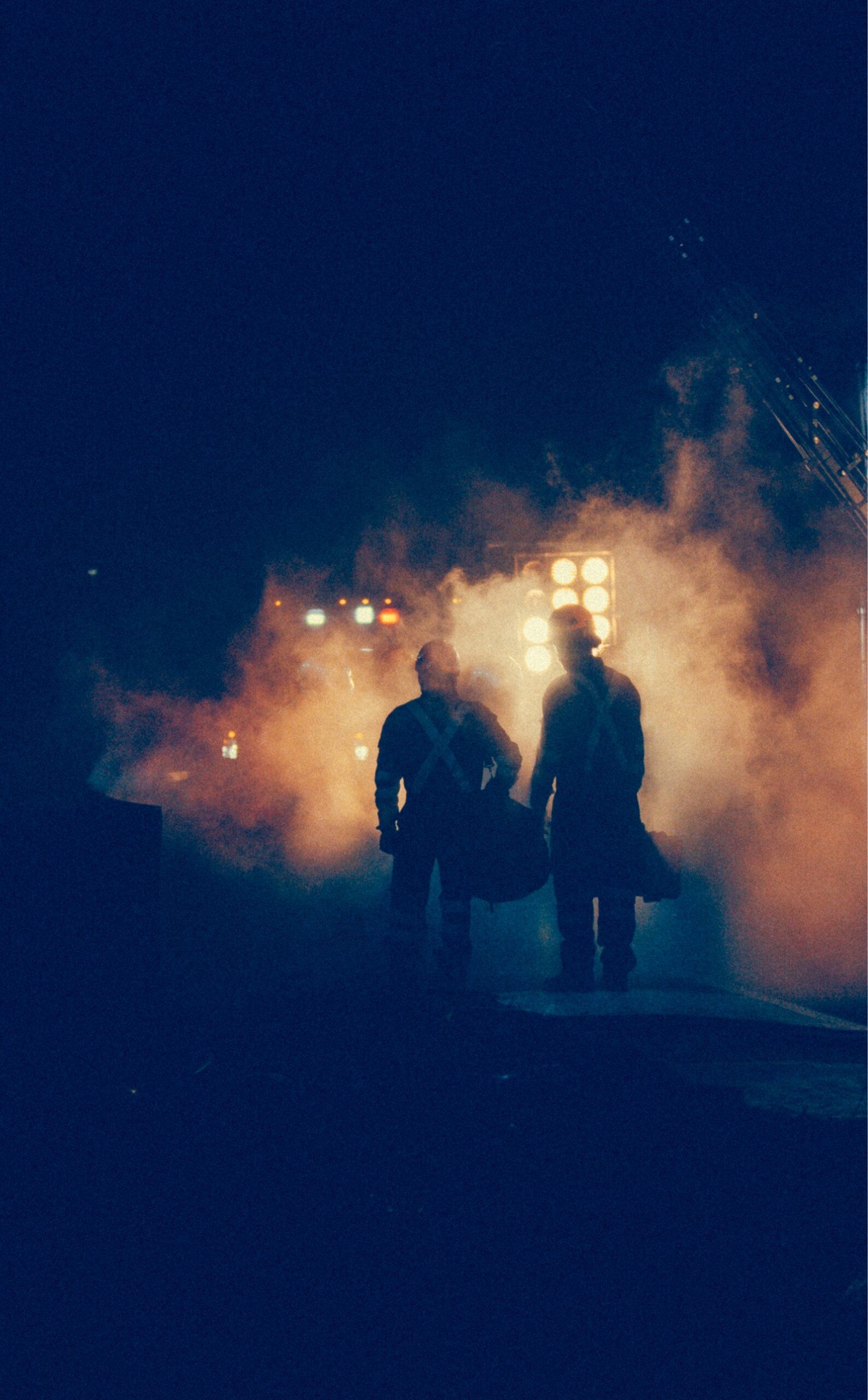 Two people in helmets and gear walk through smoke with bright lights in the background, creating a dramatic silhouette effect.