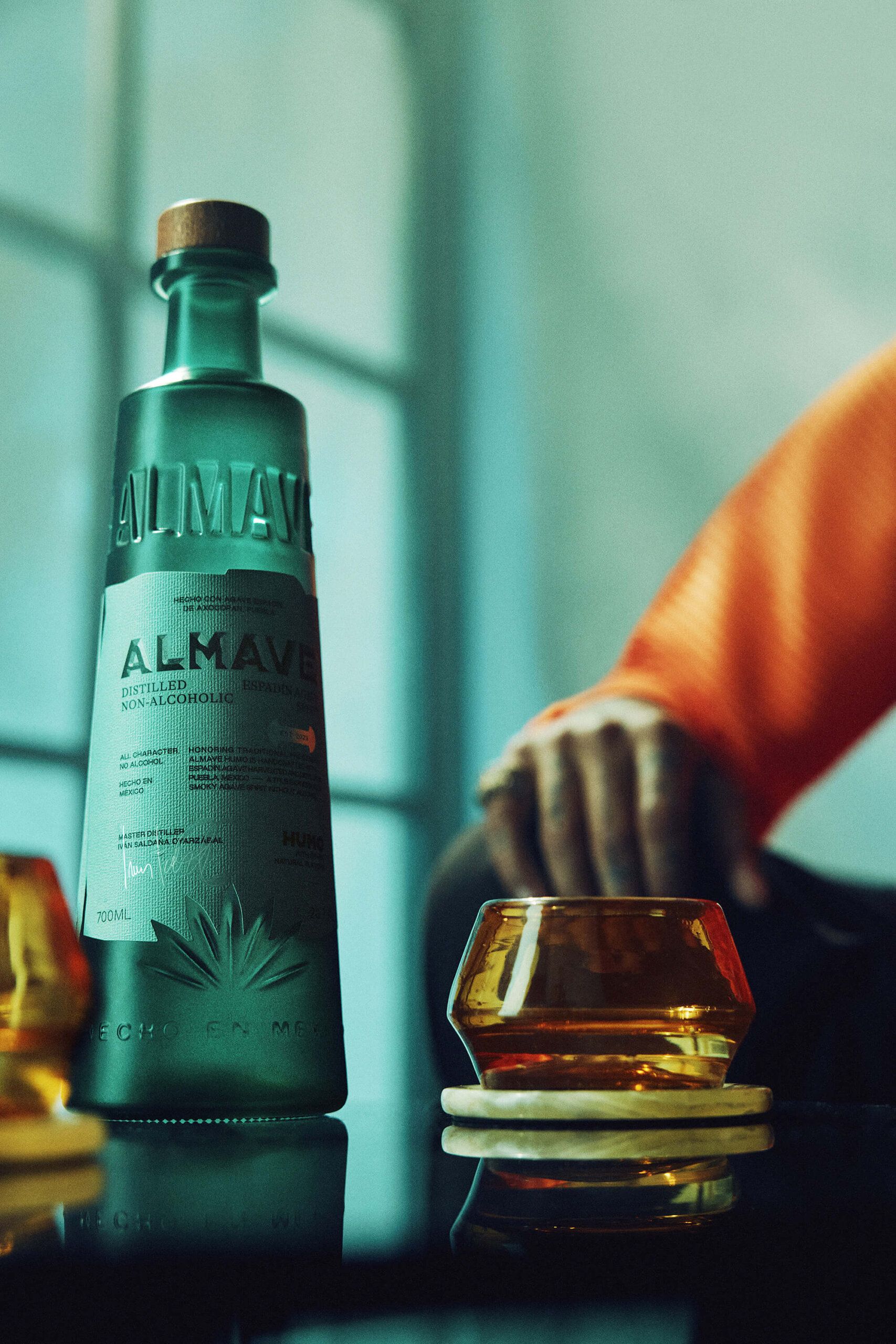 A bottle of Almave non-alcoholic spirit sits on a table next to a glass of amber liquid and a person's hand resting nearby.