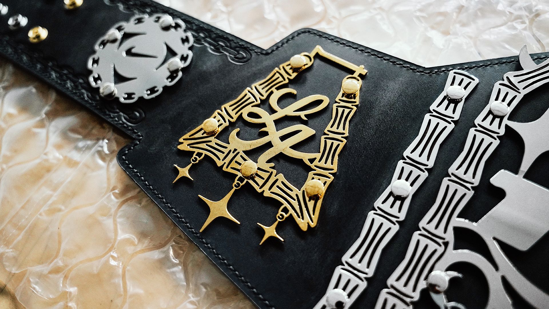 A close-up view of a championship belt with gold and silver decorative elements on a black leather background, placed on bubble wrap.