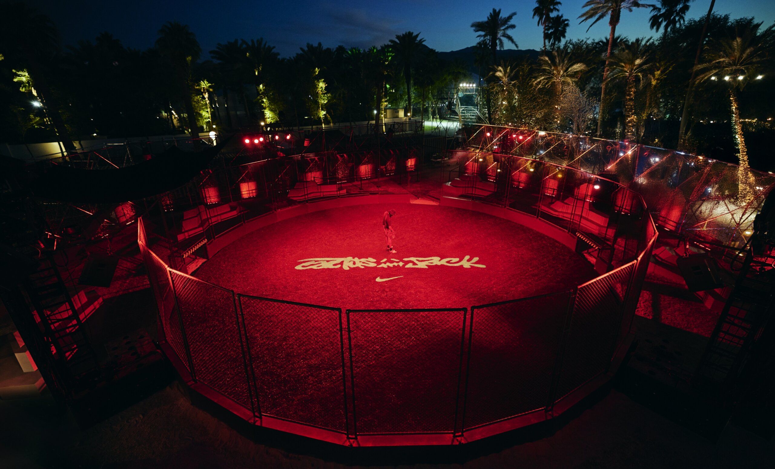 A person stands alone in the center of a circular, fenced arena illuminated with red lighting and surrounded by palm trees at night. The words "Cactus Jack" are projected onto the ground.