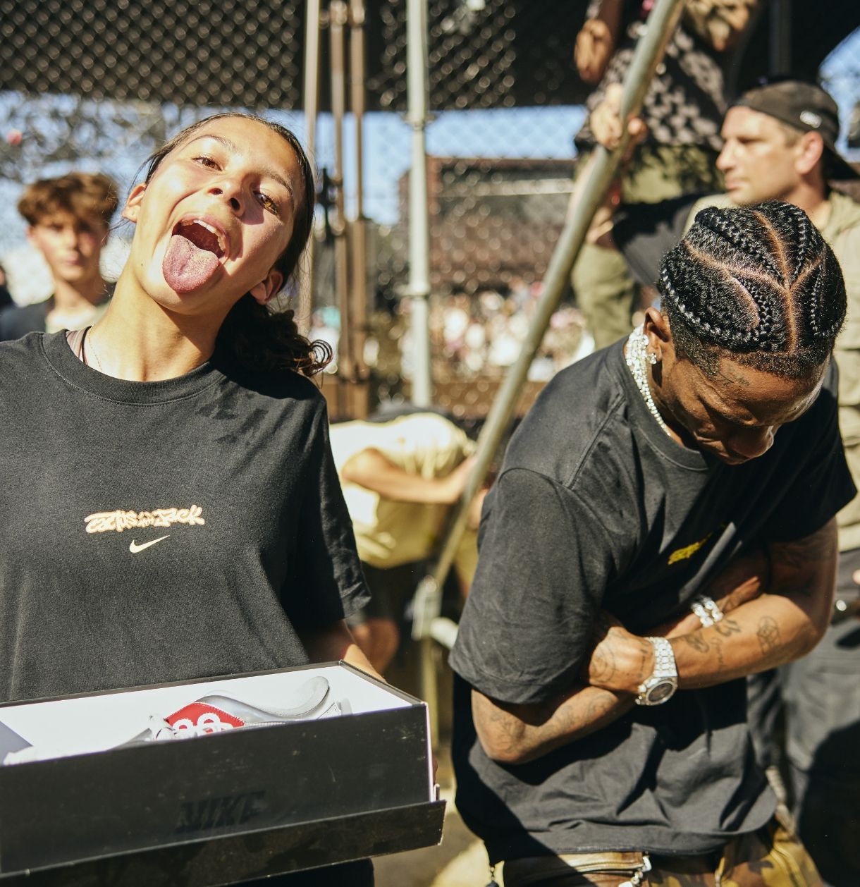 A young woman holds a Nike shoe box and sticks out her tongue while a man stands beside her with braided hair, arms crossed, at an outdoor event.