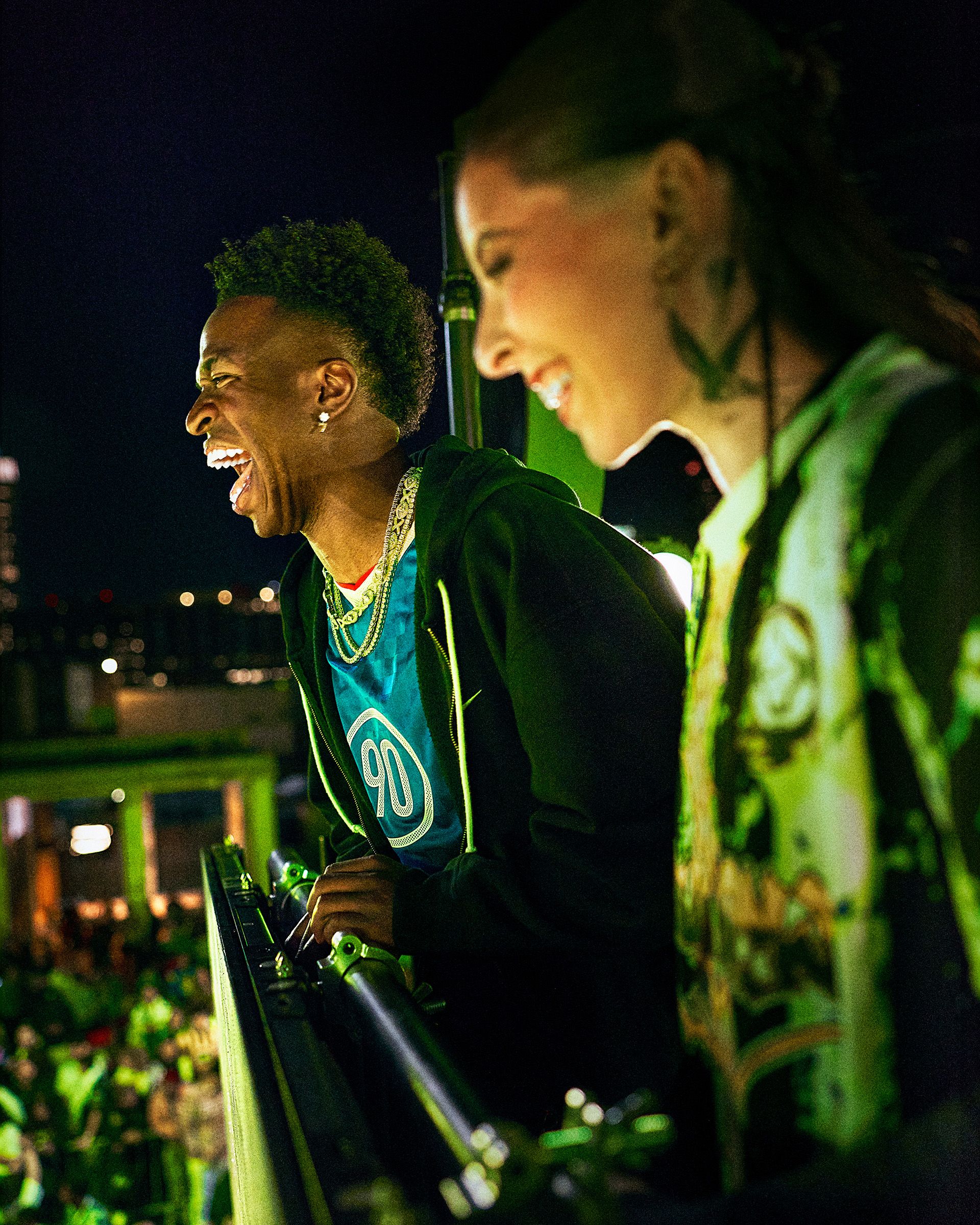 Two people stand on a balcony at night, smiling and looking down at a lively, brightly lit crowd below.