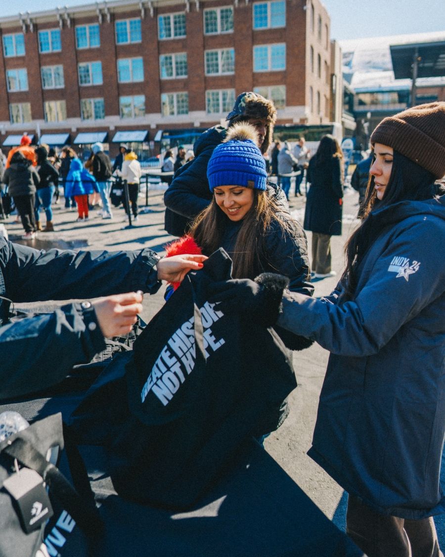 Person at table giving away tote bags to a couple people. There is a crowd of people and a large building in the background