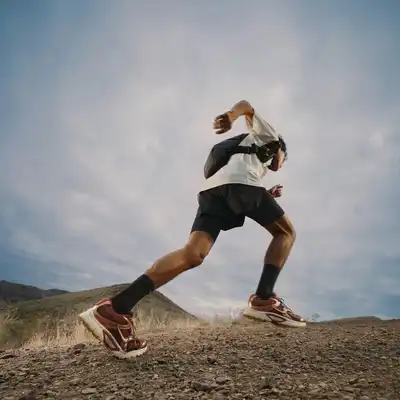 A person wearing athletic gear and a backpack runs uphill on a rocky trail with mountains and a cloudy sky in the background.
