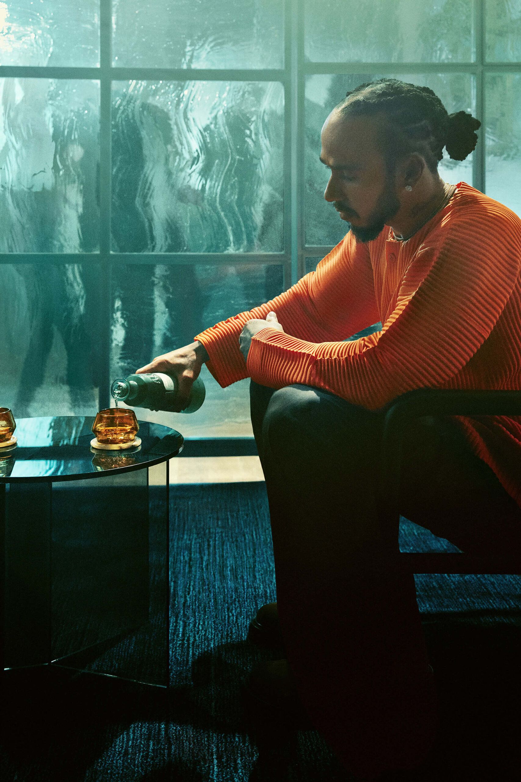 A man in an orange sweater sits in a chair, pouring a drink into a glass on a glass table, with large windows and blurred figures in the background.