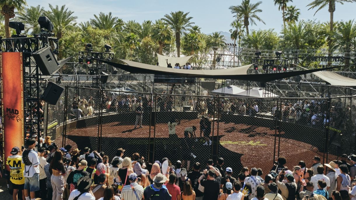 A large crowd surrounds a fenced outdoor circular stage with people inside, set among palm trees under a sunny sky.