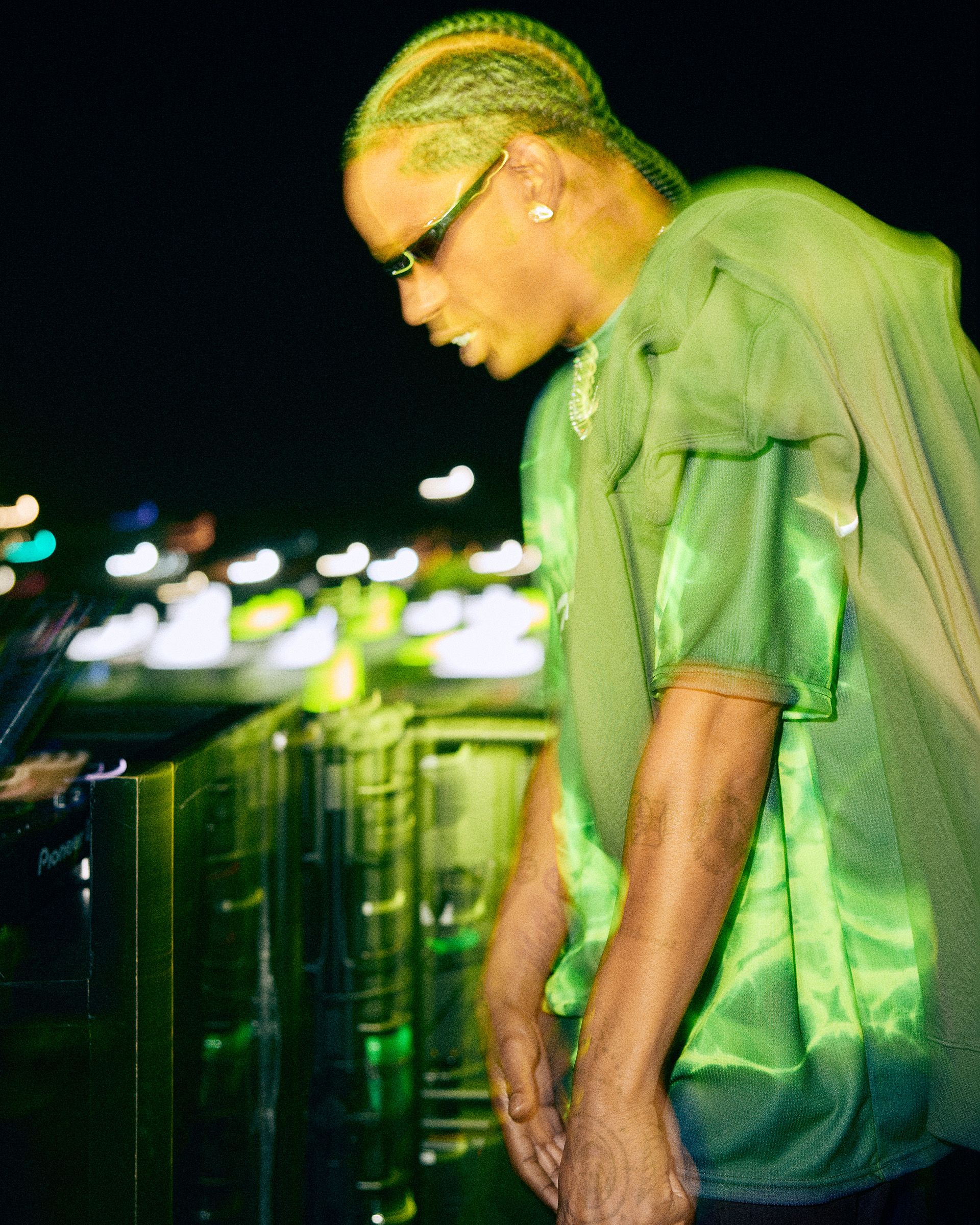 A person with braids and sunglasses, wearing a green shirt, stands outdoors at night near a glass railing with blurred city lights in the background.