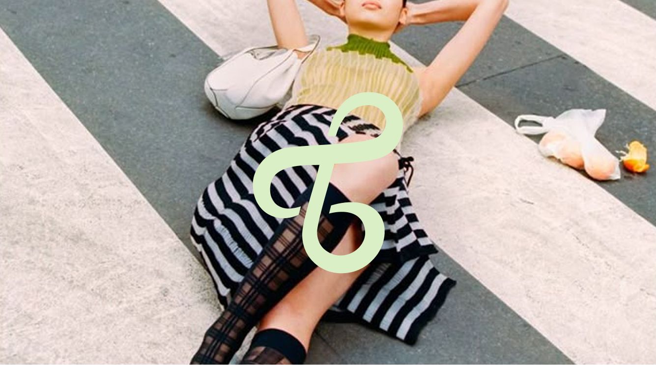 A person wearing a striped skirt and sleeveless top lies on a crosswalk, with a handbag and a plastic bag containing fruit nearby.
