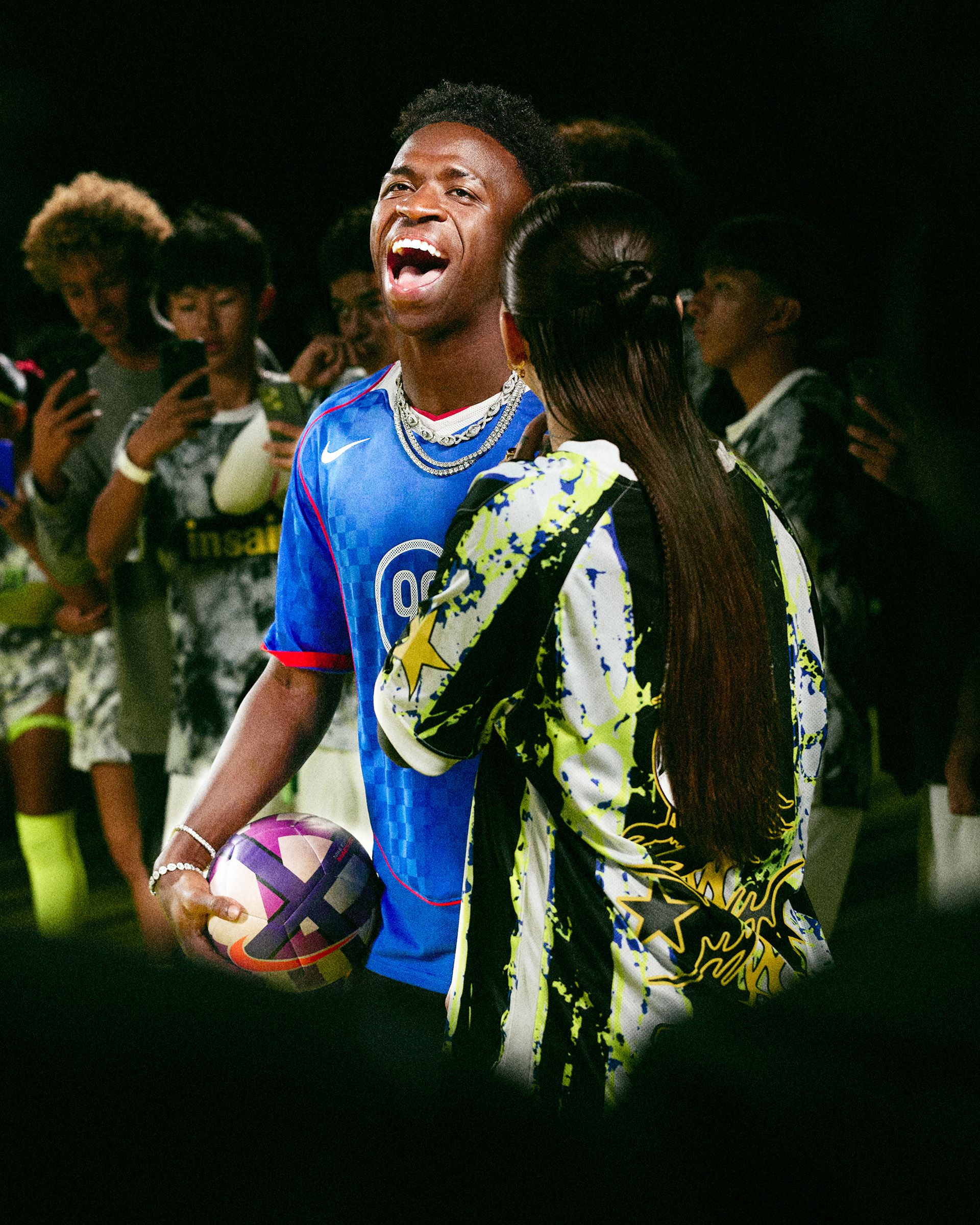 A soccer player in a blue jersey holds a ball and laughs while a woman in a patterned jacket stands beside him; people in the background take photos with their phones.