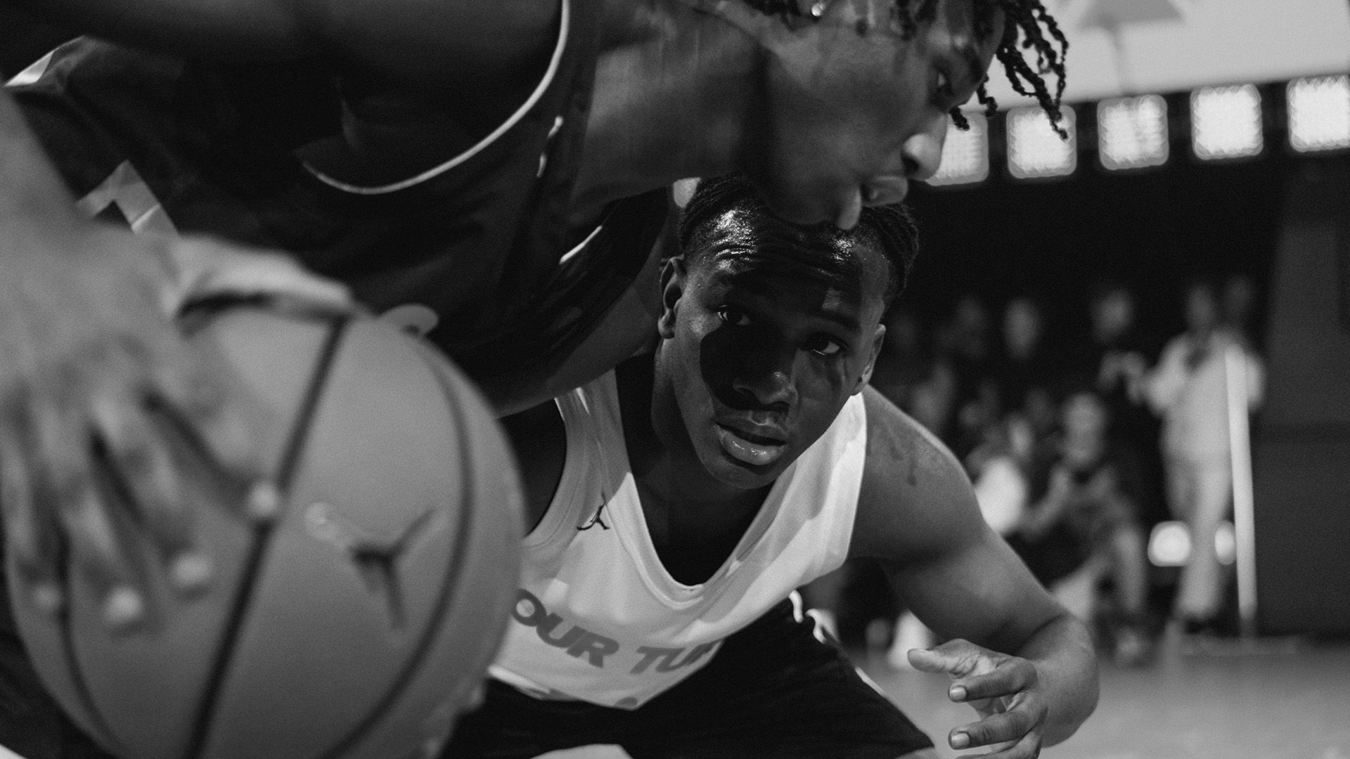 Two basketball players closely face off during a game, one dribbling the ball while the other defends; spectators are blurred in the background.
