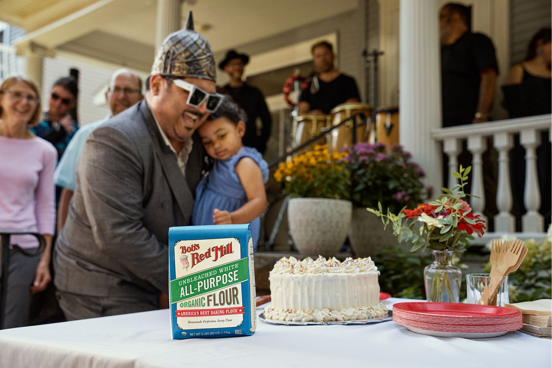 A man holding a young girl stands behind a table with a decorated cake, Bob’s Red Mill flour, and plates at an outdoor gathering in front of a house.
