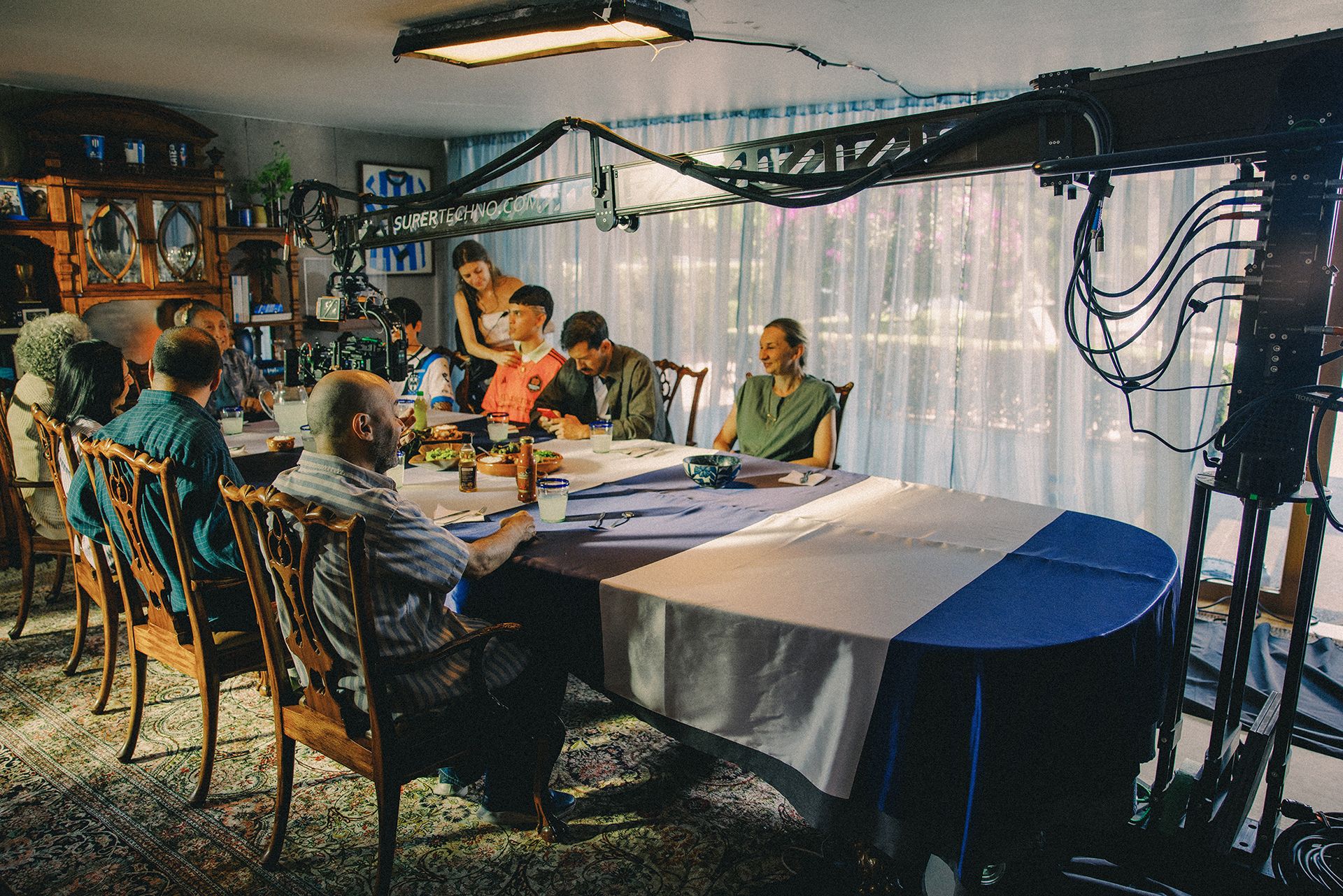 A camera crew films a group of people seated at a dining table, the table is partially set with plates and utensils. A person operates the camera mounted on a crane while others are engaged in conversation.