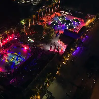 Aerial view of a brightly lit outdoor skate park event at night, surrounded by palm trees and crowds, with colorful lights illuminating the scene.