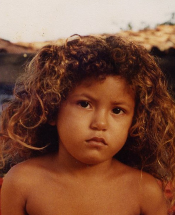 A young child with curly hair looks at the camera with a neutral expression, sitting outdoors with a tiled roof visible in the background.