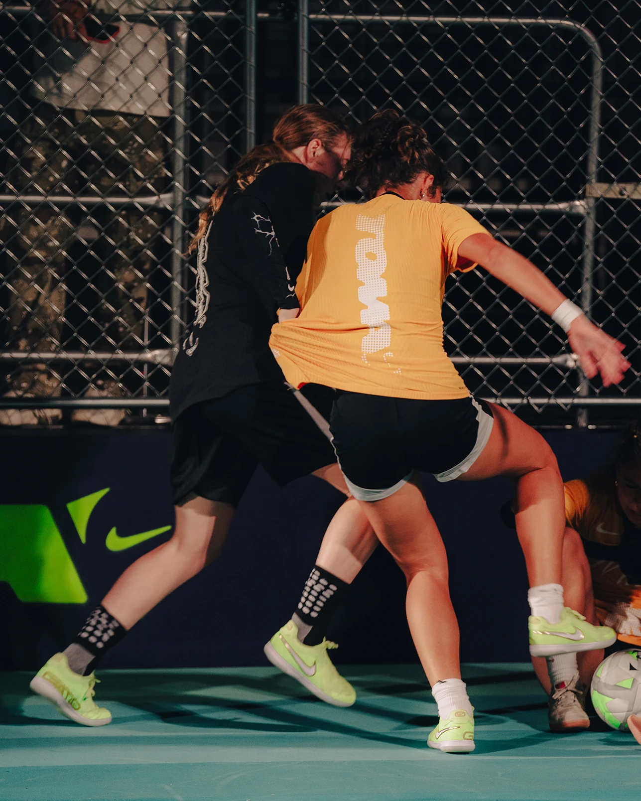Two soccer players compete for the ball on an indoor court, with one in a yellow shirt and the other in black, near a metal fence background.