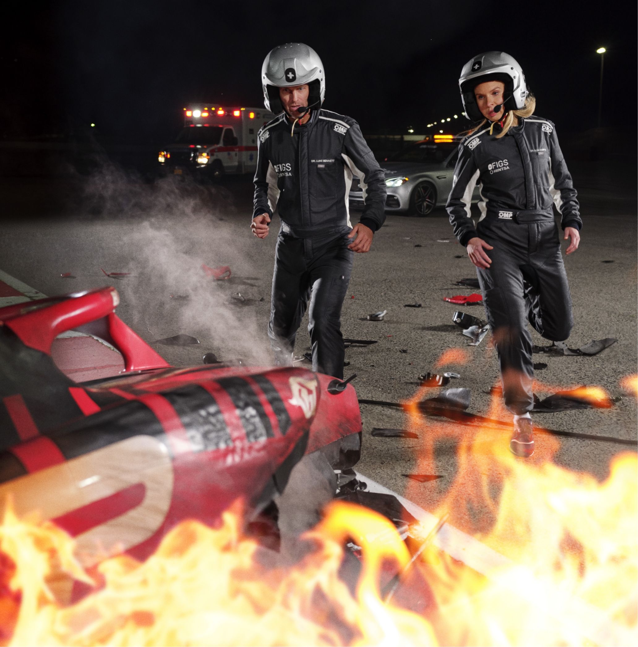 Two people in racing suits and helmets walk toward a burning race car surrounded by debris, with an emergency vehicle in the background.