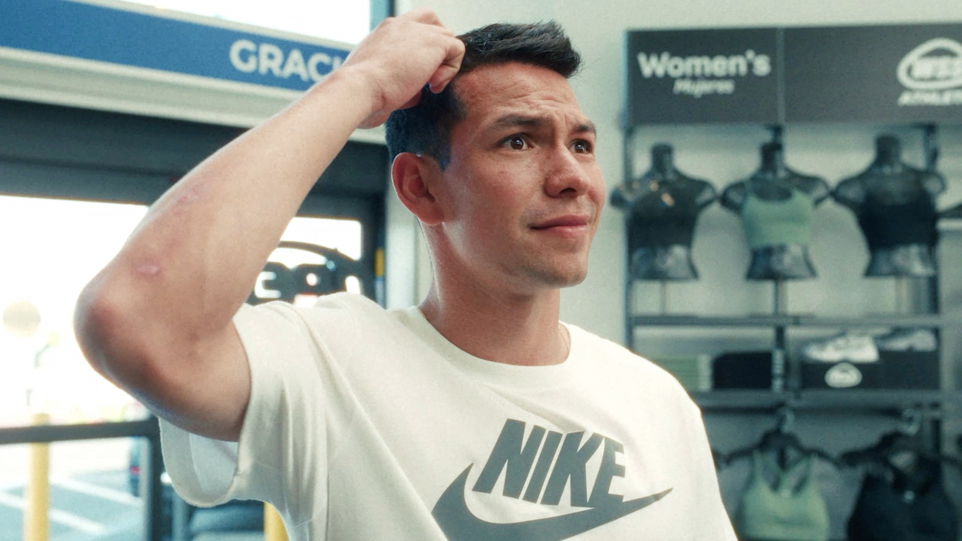 A man in a white Nike t-shirt stands indoors, scratching his head with a confused expression; sports mannequins and signs are visible in the background.