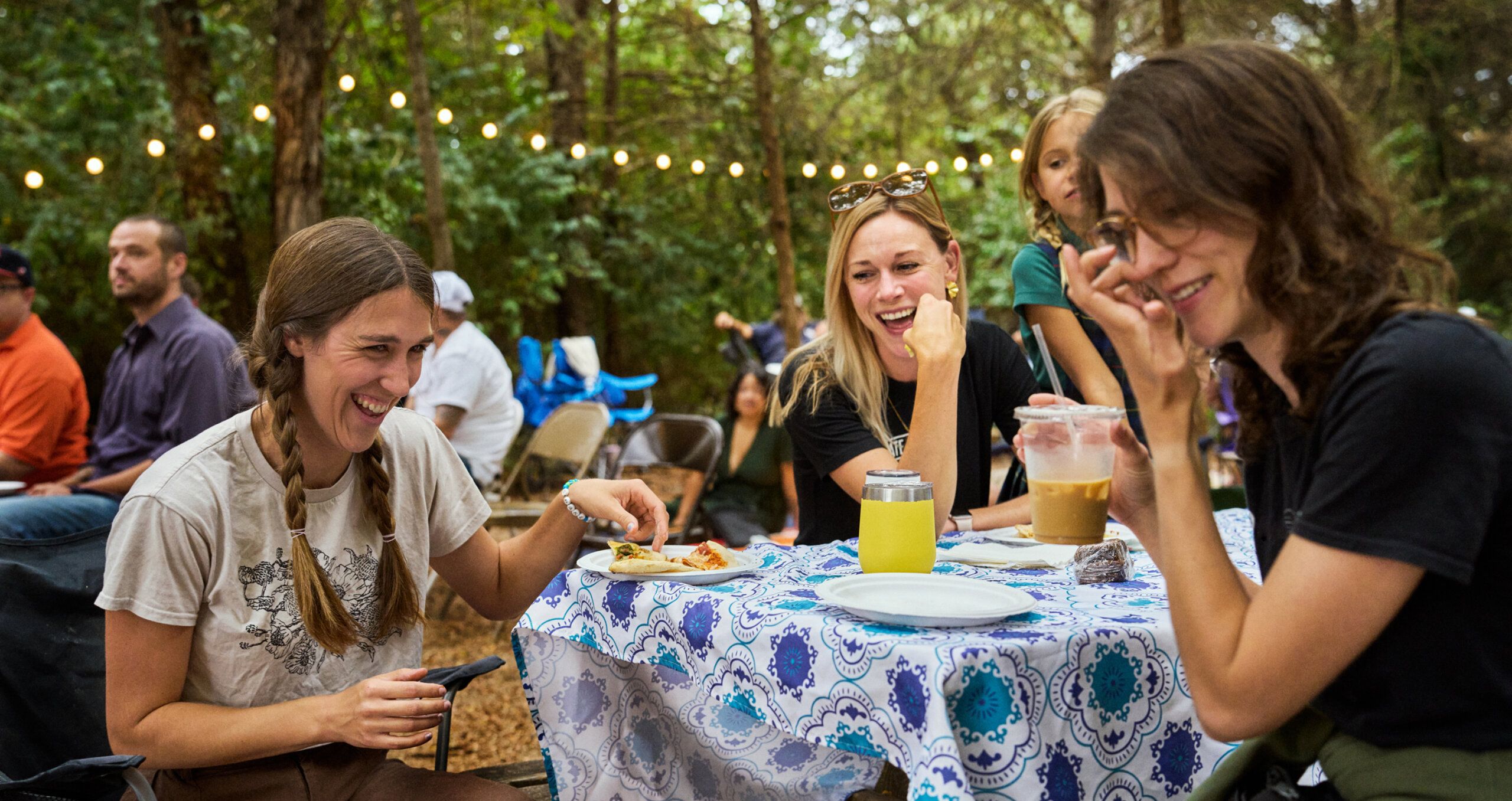 Three women sit at a table outdoors, laughing and eating, with string lights and people in the background in a wooded area.