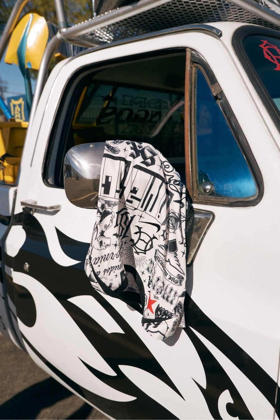 A white truck with black flame decals has a graphic black and white shirt draped over its side mirror.