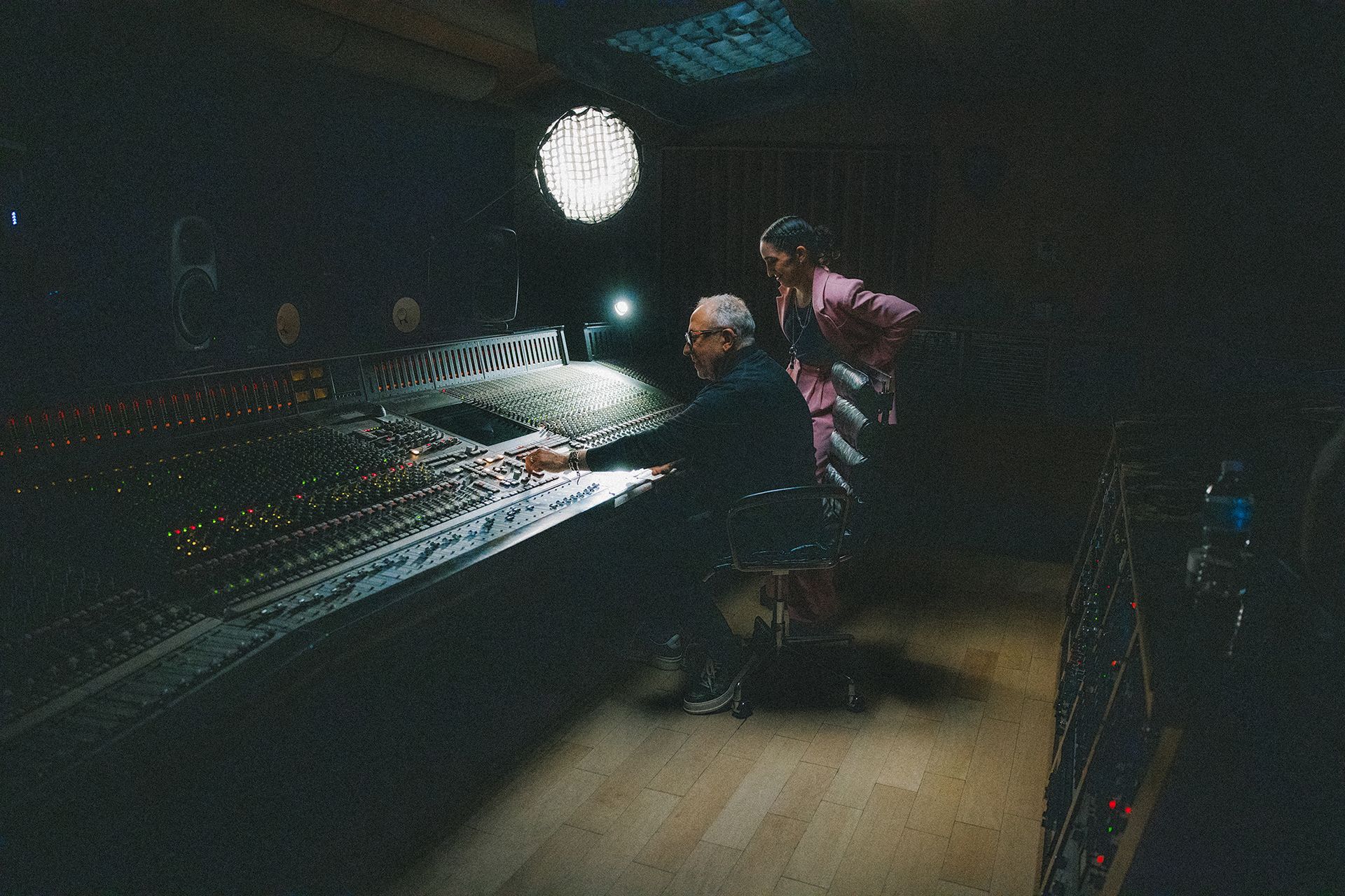 Two people are working in a dimly lit recording studio. One is seated at a large mixing console, and the other stands behind, observing.