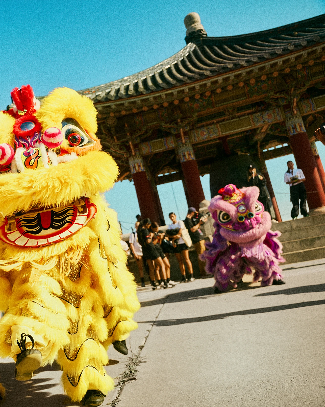 Two performers in bright yellow and purple lion dance costumes perform near a traditional Asian pavilion, with onlookers and clear blue sky in the background.
