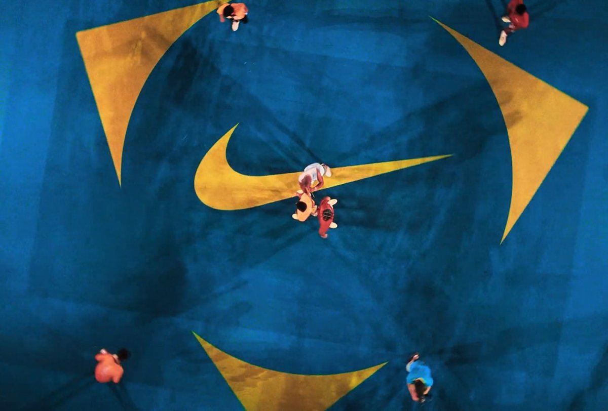 Aerial view of six people standing and moving on a blue sports court with large yellow Nike logos painted on the surface.