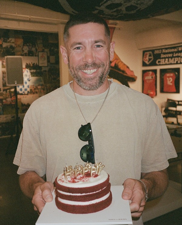 A smiling man in a beige shirt holds a red velvet birthday cake with candles that spell "HAPPY BIRTHDAY" in a room decorated with sports memorabilia.