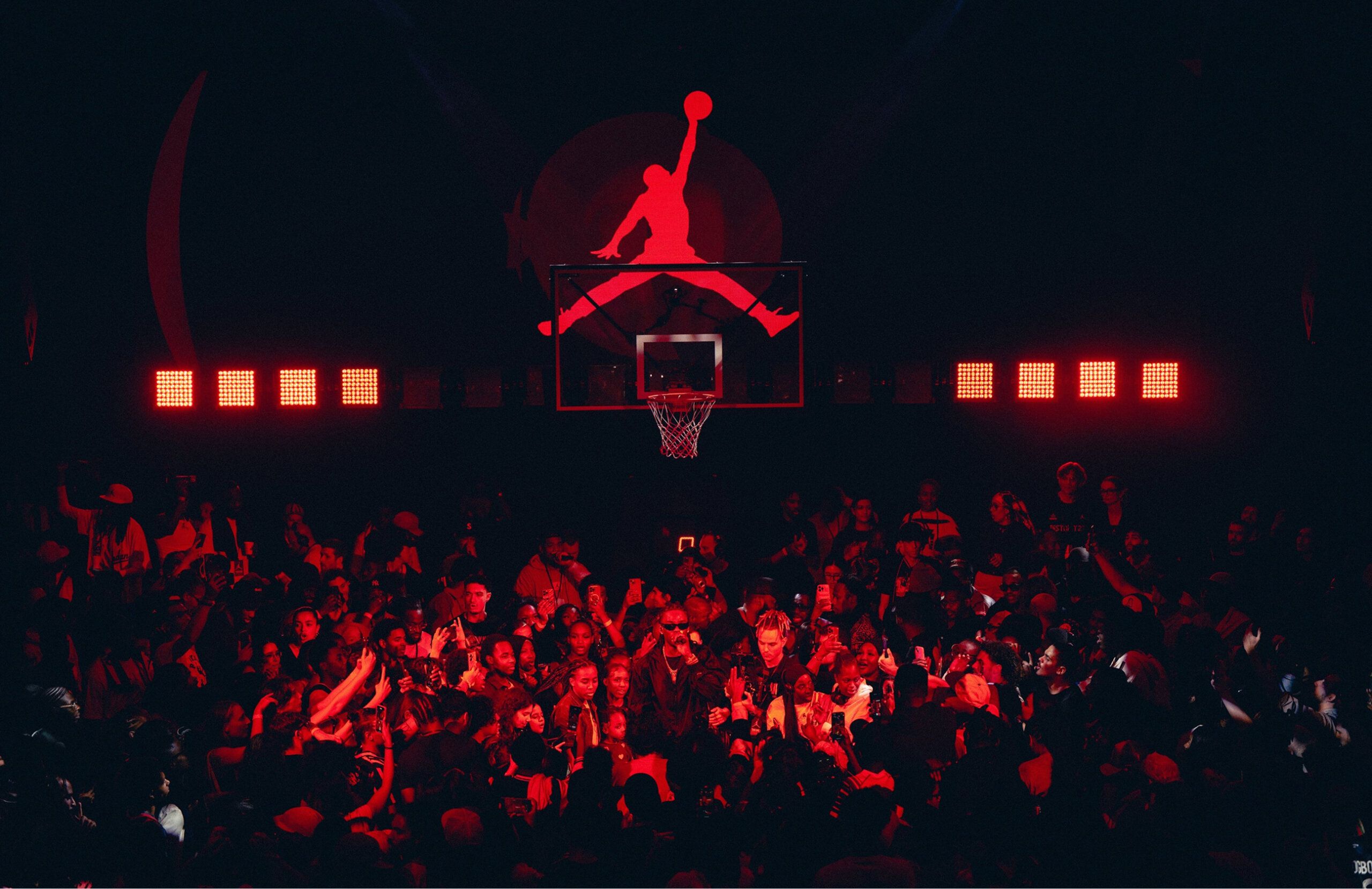 A crowd gathers beneath a basketball hoop at night, illuminated by red lighting. A large Jumpman logo is displayed above the hoop.