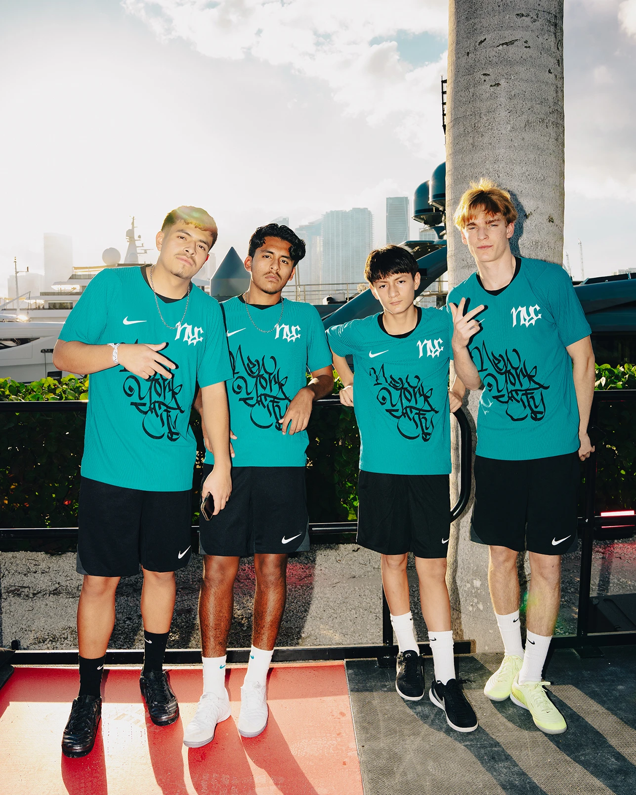 Four young men wearing matching teal shirts and black shorts stand together outdoors, posing for a group photo with a city skyline in the background.