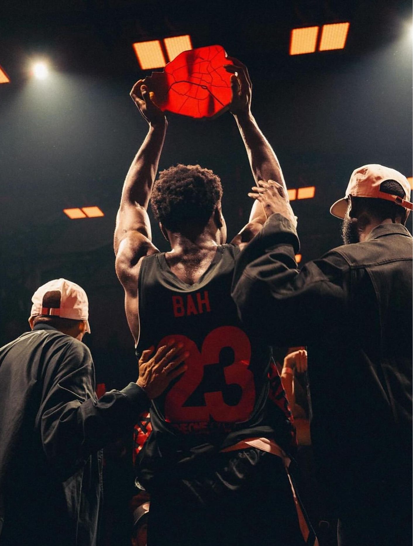 A basketball player wearing a jersey labeled "BAH 23" holds a red item above his head while two others in hats stand beside him, under bright indoor lights.