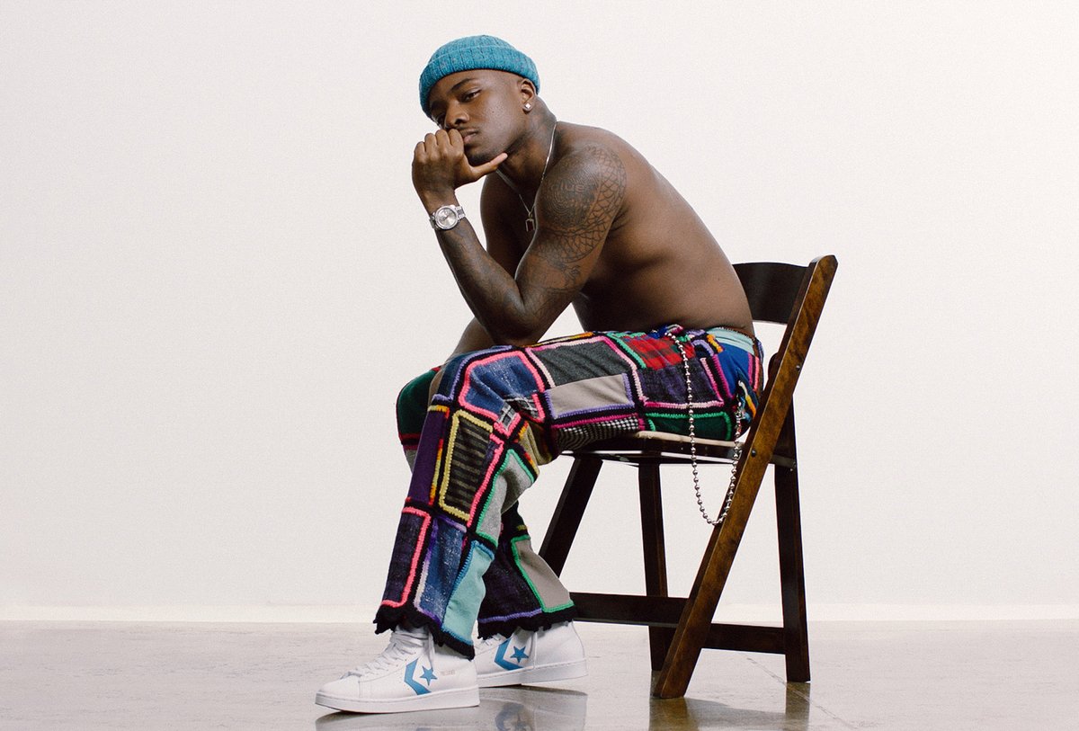 A shirtless man wearing a blue beanie, patchwork pants, and white sneakers sits sideways on a wooden chair against a plain background.
