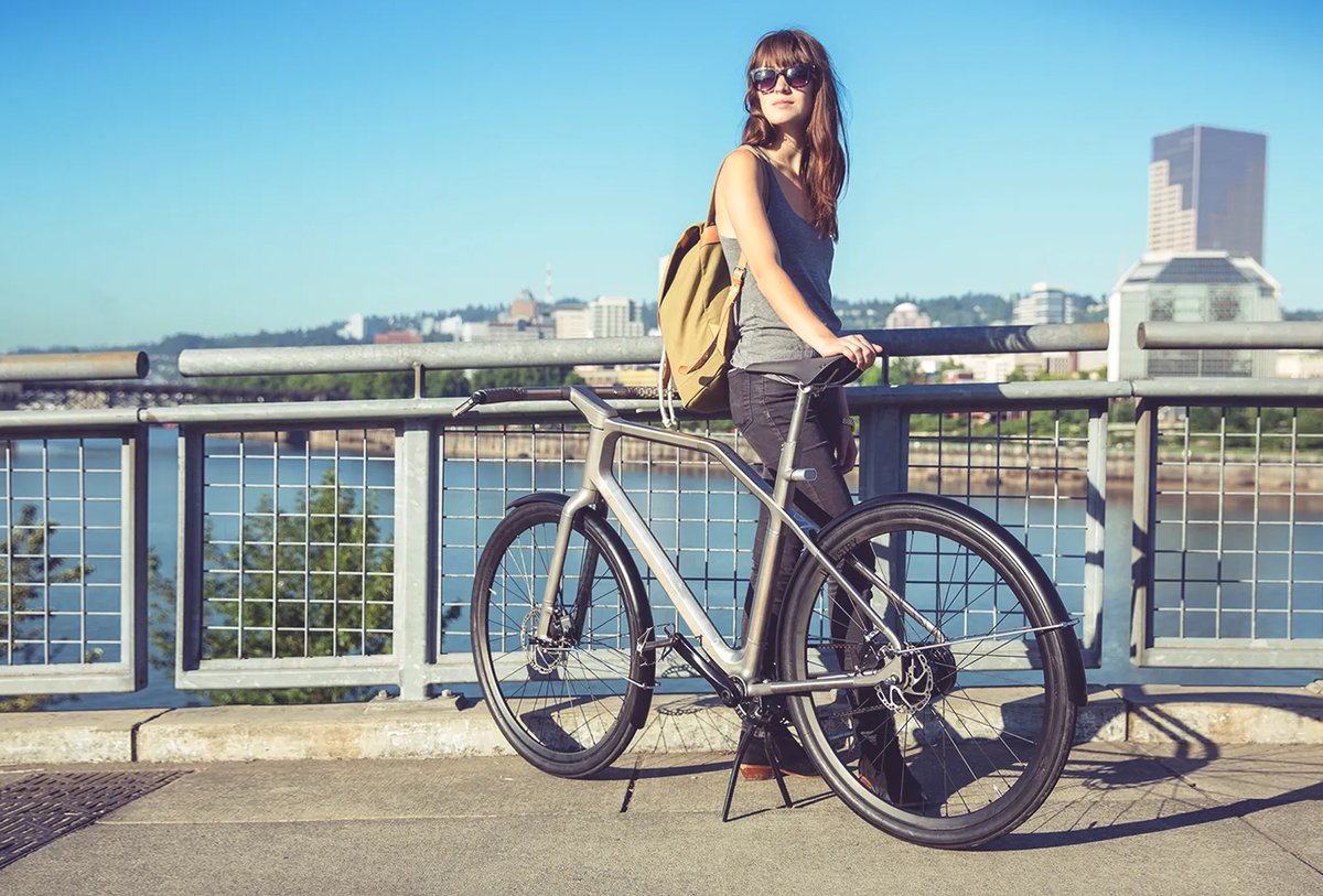 A woman wearing sunglasses and a backpack stands next to a bicycle on a bridge, with a cityscape and river in the background.