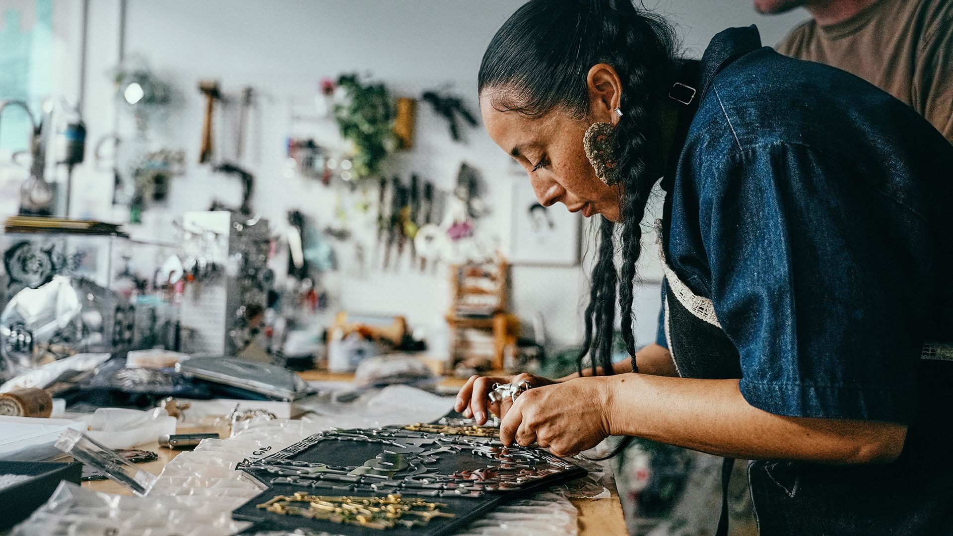 A woman with braided hair examines and arranges handcrafted jewelry pieces on a table in a workshop filled with tools and materials.