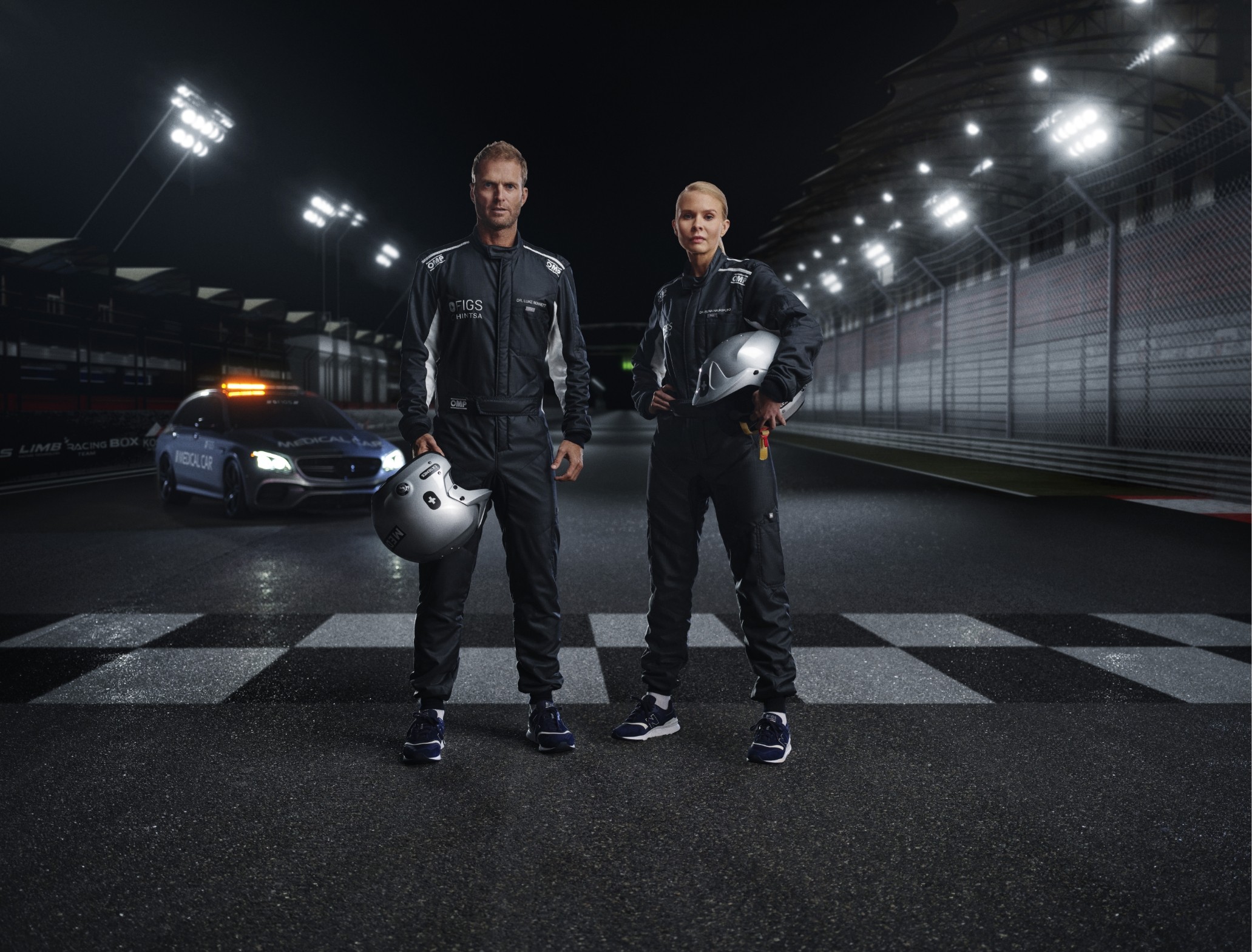 Two people in racing suits stand on a wet track. One holds a helmet. A safety car with flashing lights is in the background.