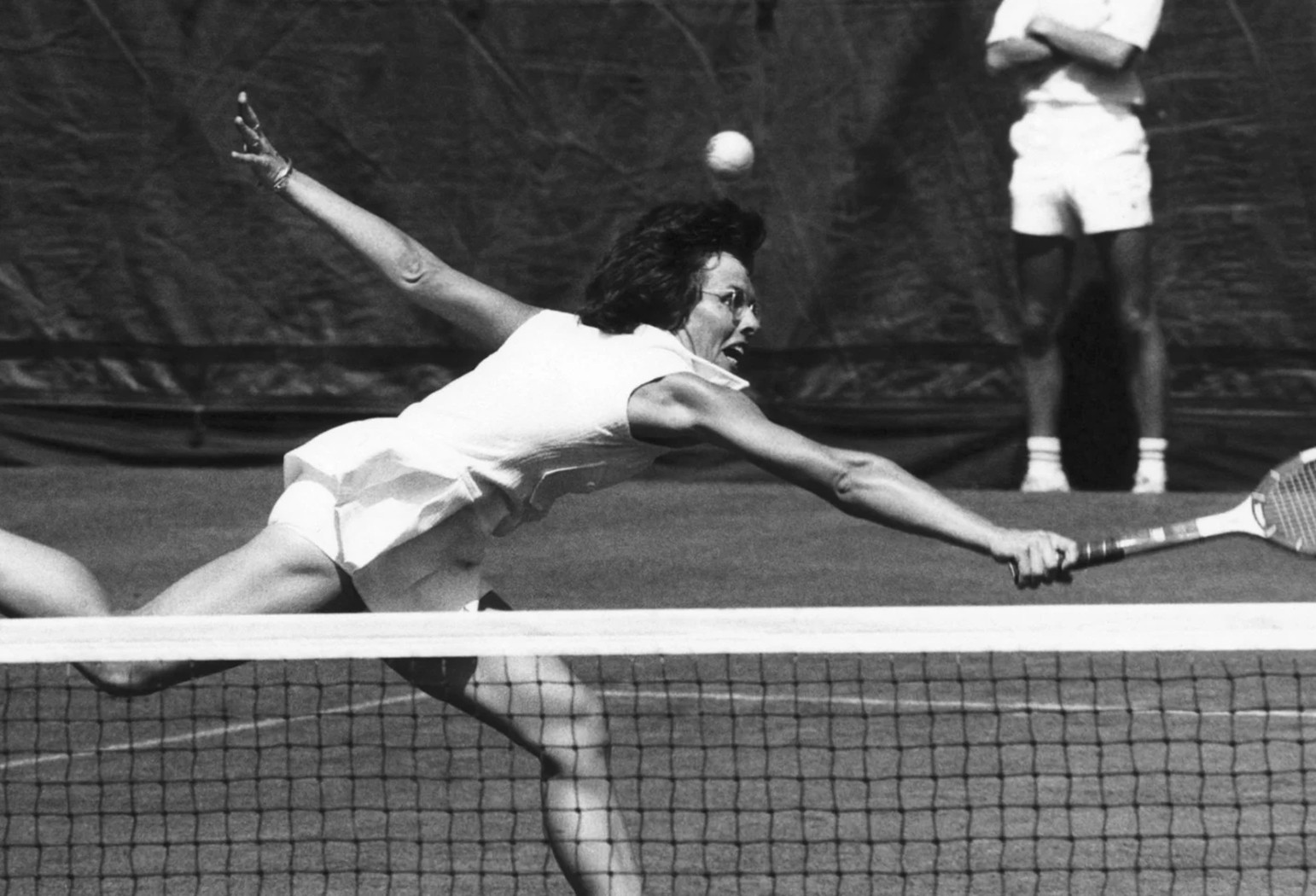 A tennis player lunges forward to hit a forehand volley at the net during a match on a grass court.