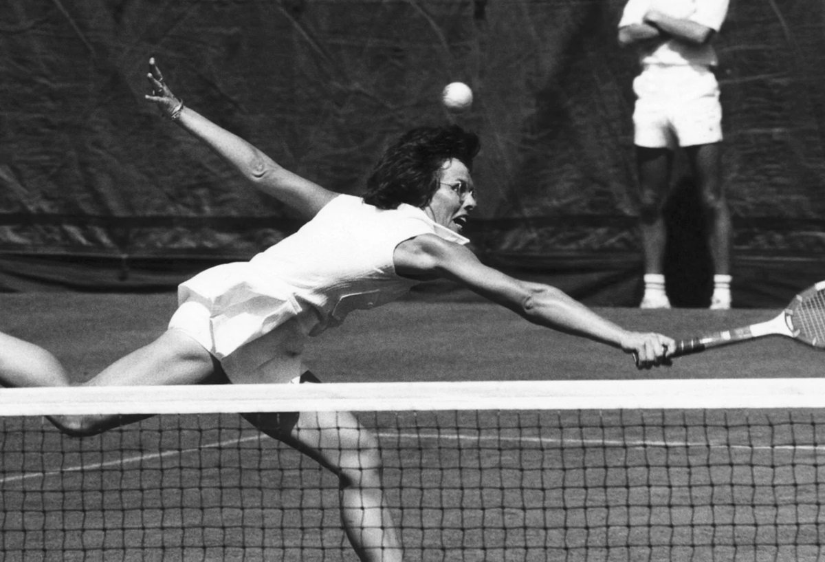 A tennis player lunges forward to hit a forehand volley at the net during a match on a grass court.