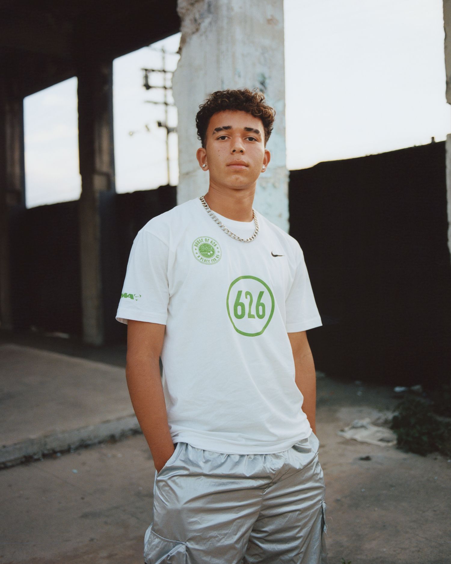 A young man stands outdoors wearing a white t-shirt with green graphics and silver shorts, hands in pockets, looking at the camera. The background includes concrete pillars and a black fence.