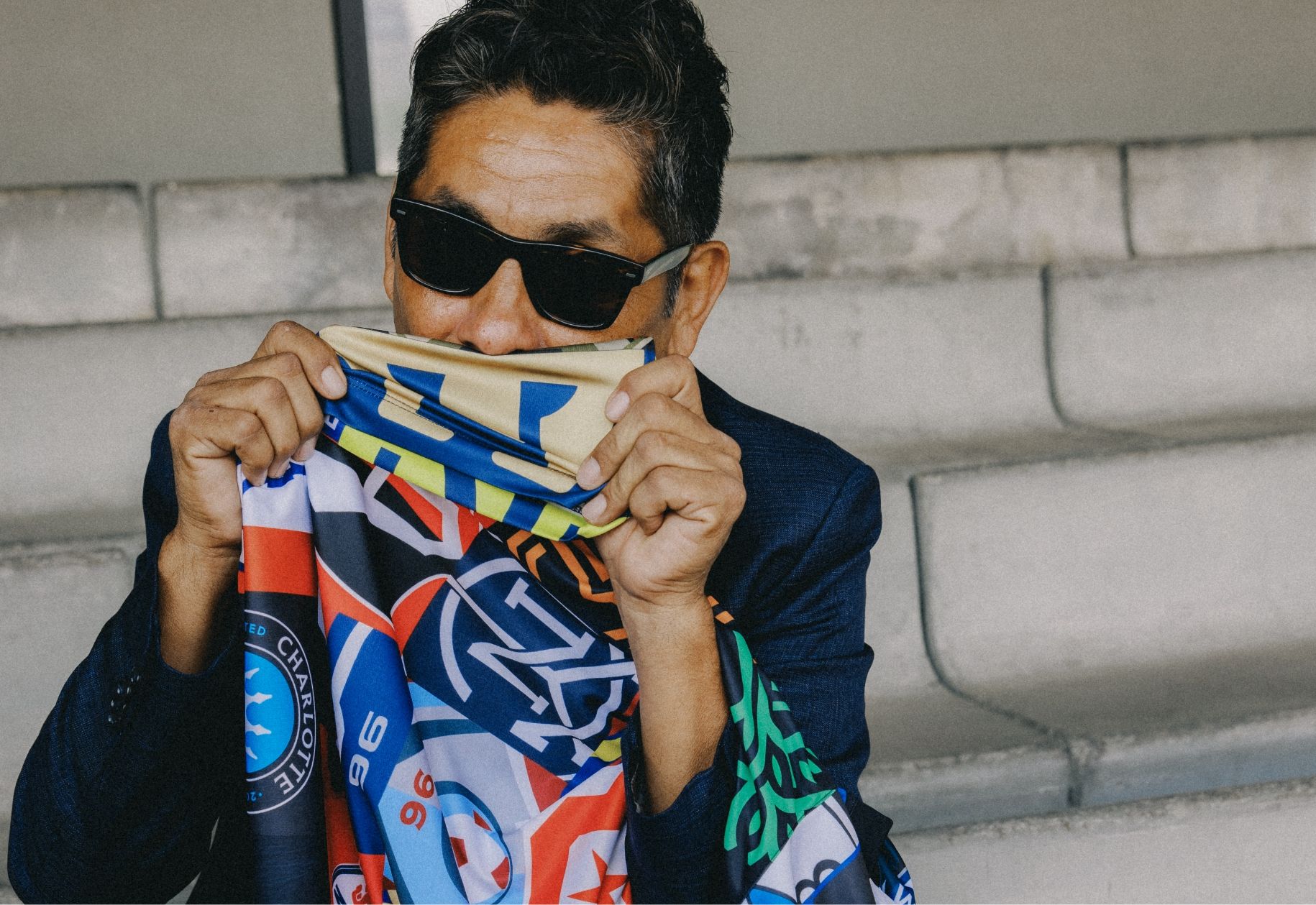 A person wearing sunglasses and a dark suit holds up a colorful patterned fabric covering their mouth while sitting on concrete steps.