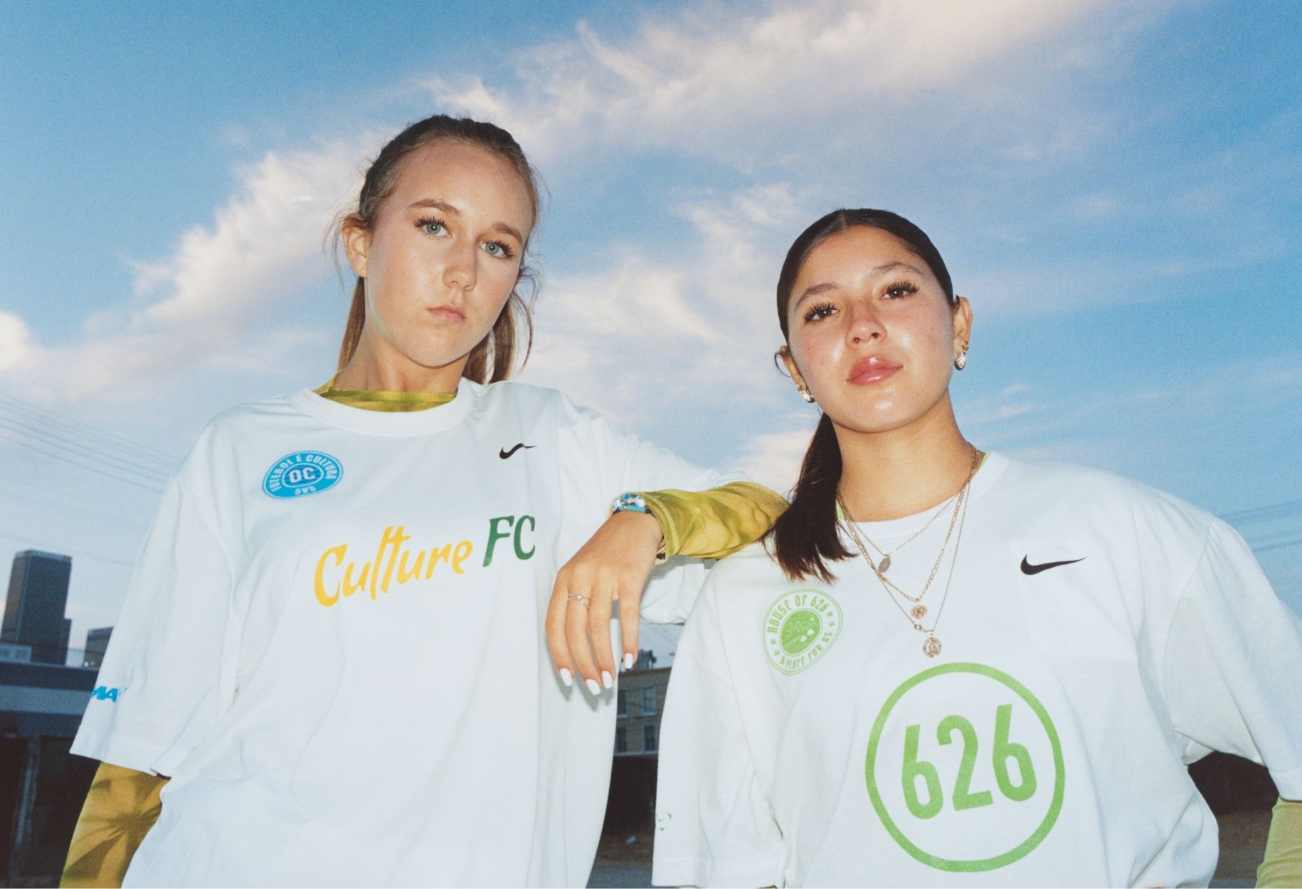 Two women stand side by side wearing white Culture FC jerseys with logos and numbers, looking at the camera against a blue sky background.