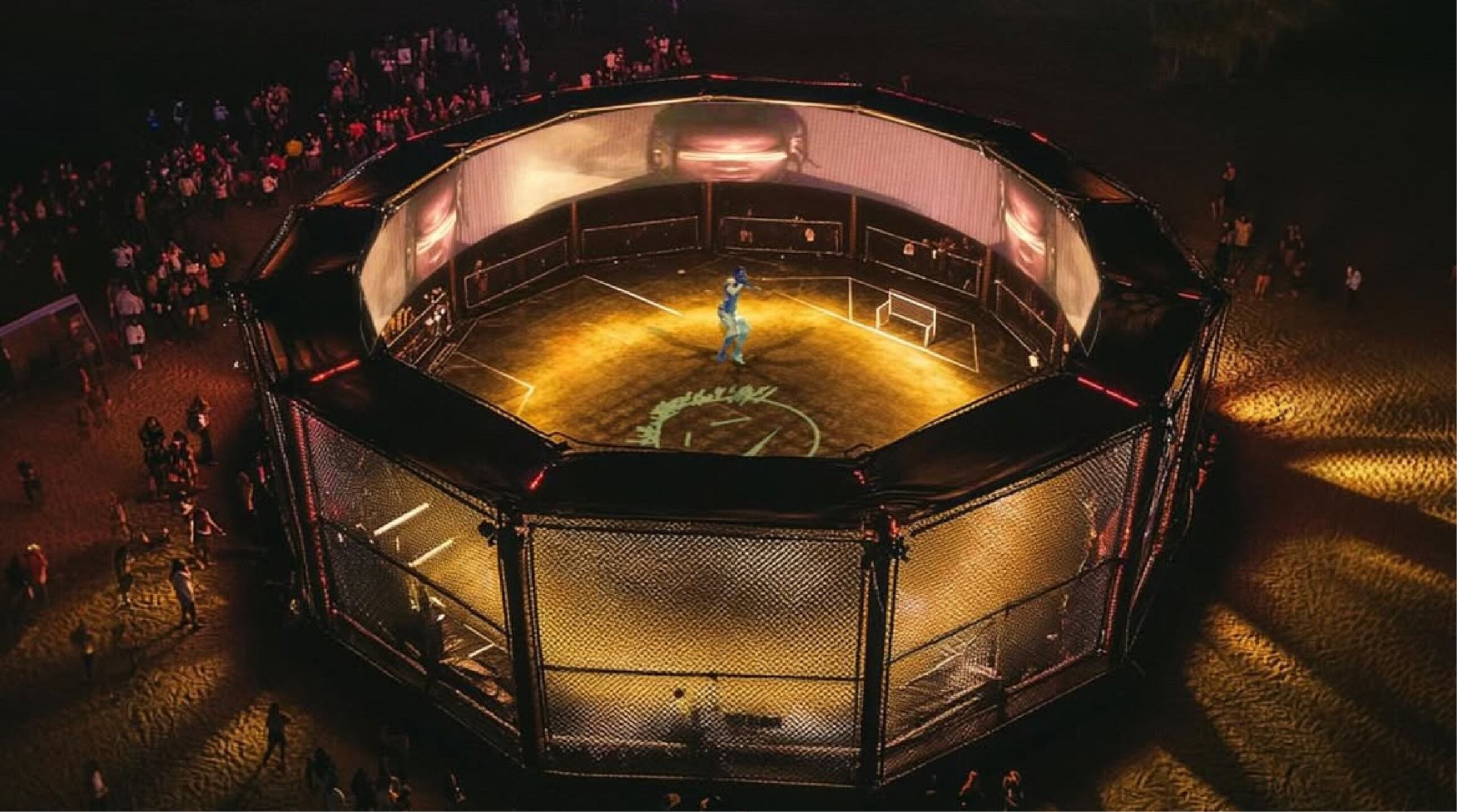 Aerial view of a brightly lit, circular outdoor sports arena at night with one person inside and a crowd gathered around the perimeter.