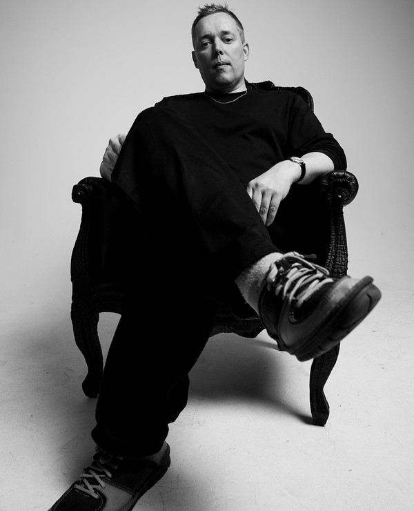 A person sits in a large armchair with legs crossed, wearing dark clothing and sneakers, looking directly at the camera in a plain studio setting.