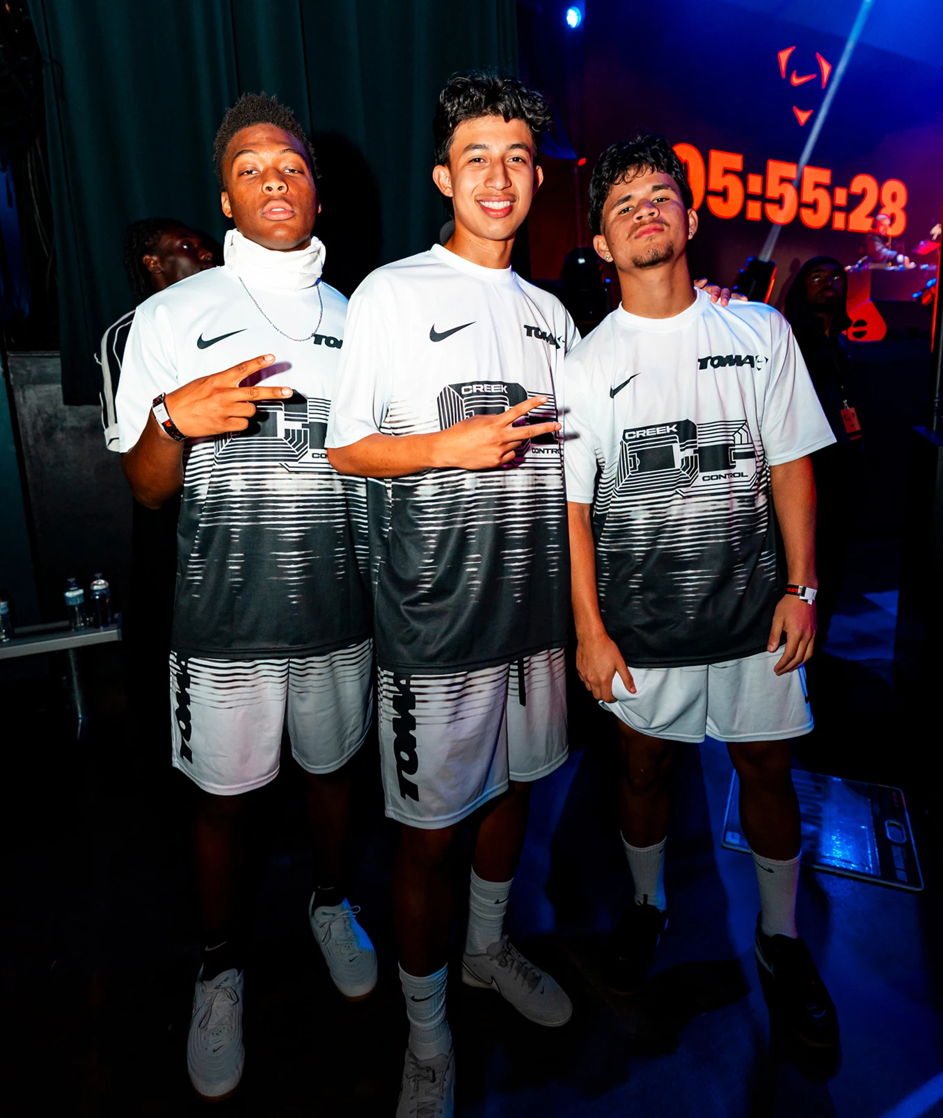 Three young men in matching black and white sportswear pose indoors, making peace signs. A large digital clock and stage lighting are visible in the background.