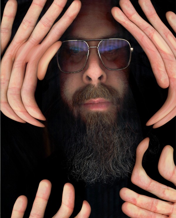 A bearded person wearing glasses presses their face and multiple hands against a glass surface, creating a distorted reflection effect.