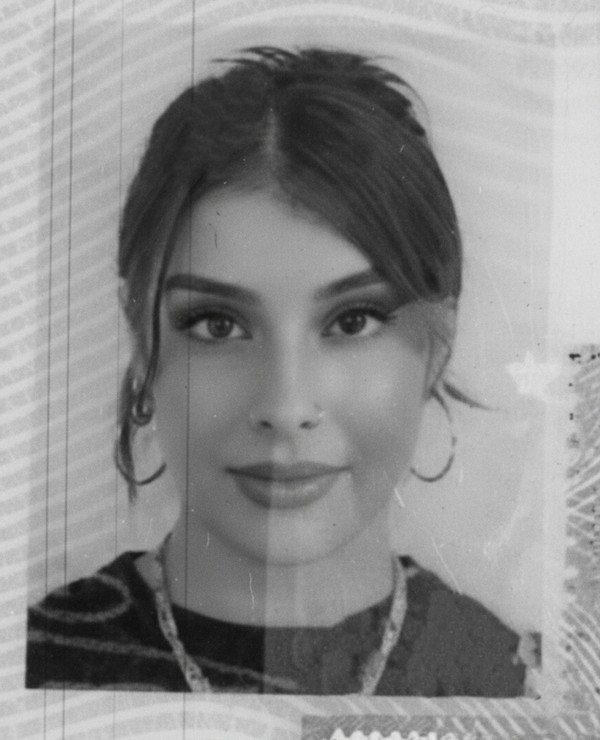 Black and white headshot of a young woman with long hair pulled back, wearing hoop earrings, a necklace, and a dark top, looking straight at the camera.