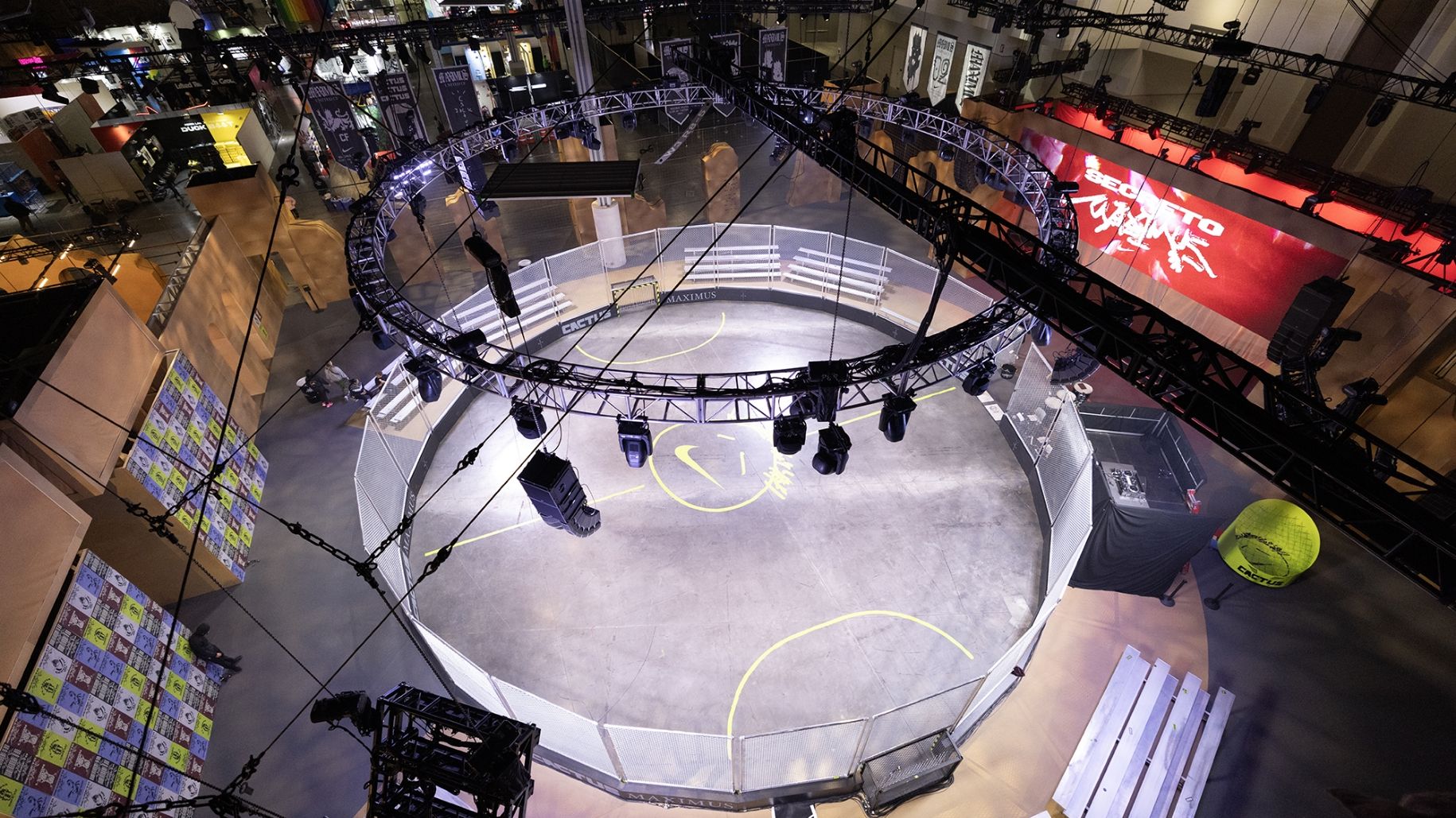 A circular gaming arena with overhead rigging and lighting, surrounded by a safety fence. Displays and booths are visible in the background.